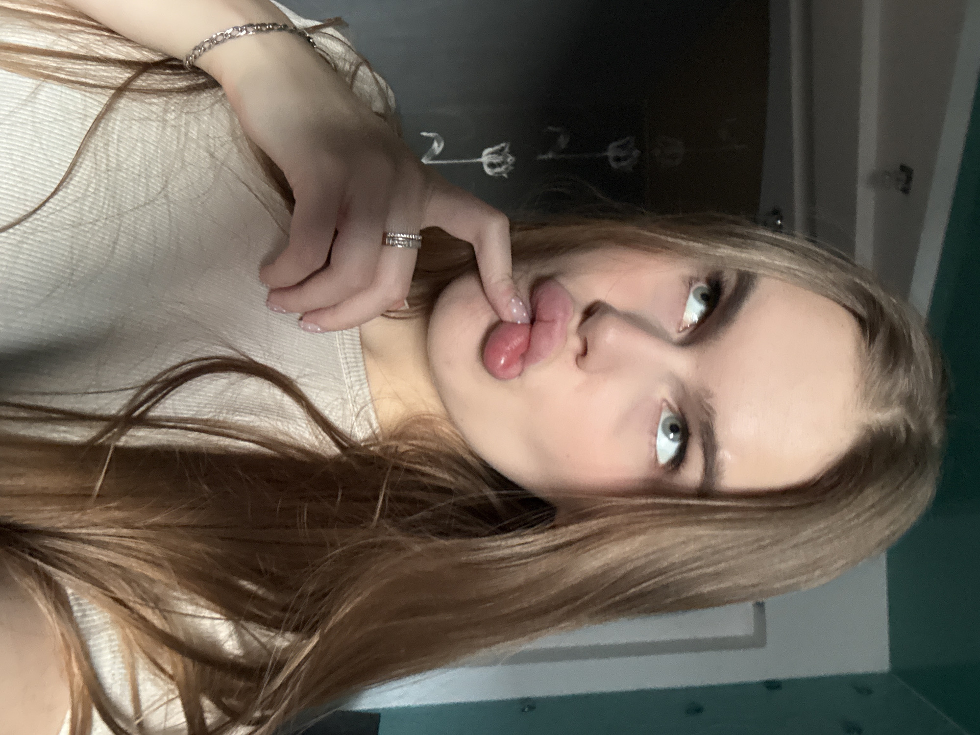 The media file shows a close-up, portrait-oriented selfie of a young woman with long, light brown hair. She is wearing a light-colored, ribbed top. On her left wrist, she wears a silver chain bracelet, and on her ring finger, she has a silver ring with a textured band. Her right hand is near her face, with her index finger gently touching her lips. She is sticking her tongue out, which is slightly curled. Her eyes, which appear to be a light blue or green, are looking towards the camera. The background is dark and appears to be an indoor setting, possibly a room. There are some faint white line drawings on the dark background, resembling stylized flowers or plants, and possibly the numbers "2022." The lighting suggests it is daytime, but the indoor setting makes it difficult to determine the exact time. There are no obvious weather cues. The overall emotion conveyed is playful or cheeky. The location context is Dnipro, Ukraine.