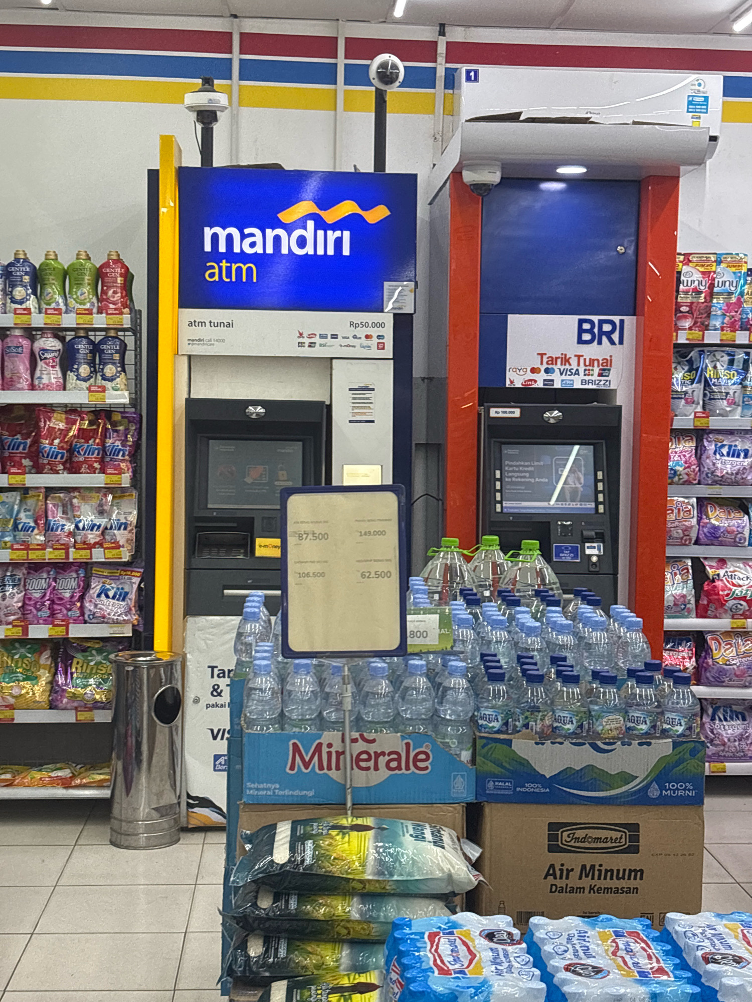 The image depicts a convenience store interior in Bekasi, Indonesia, likely an Indomaret, given the branding. Two ATMs are visible, one branded "mandiri atm" and the other "BRI Tarik Tunai." Both ATMs are functional, with screens displaying interface prompts. In front of the ATMs, there are stacks of bottled water and various packaged goods, including detergent and rice, suggesting a retail setting. A metallic trash can is positioned to the left of the ATMs. The overall environment appears to be a well-lit, organized retail space. There are no people visible in the image.