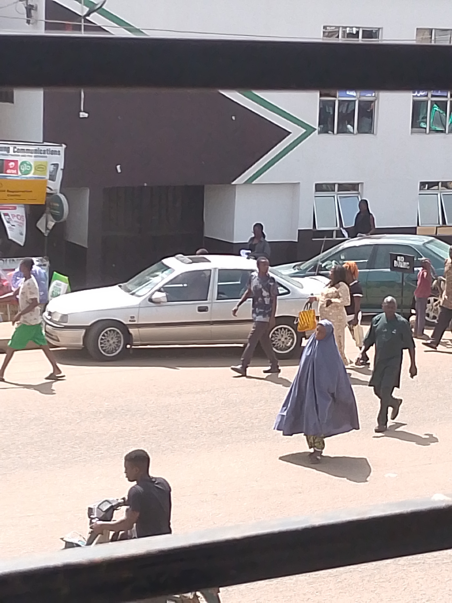 The image depicts a street scene in Bukuru, Nigeria. In the foreground, a person sits on a scooter. Further back, several pedestrians are walking. One person in a blue hijab is prominent, while others are dressed in various casual attire. Two cars are visible on the street: a silver sedan and a green car with a "NO SMOKING" sign on the rear window. The background shows a building with large windows. The overall scene is indicative of a busy urban environment with pedestrian and vehicular traffic.