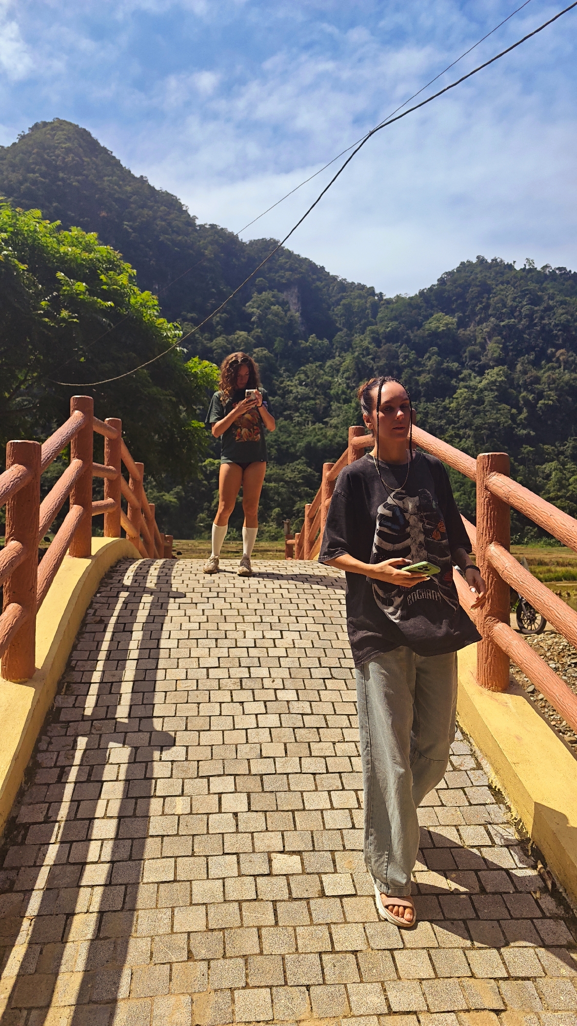 Two women are on a paved bridge in Pù Luông Commune, Vietnam. The woman in the foreground, on the right, is walking towards the viewer and wearing a black t-shirt with a graphic print, light blue pants, and white sandals. She is holding a green smartphone in her left hand. The woman in the background, on the left, is standing still and looking at her phone, wearing a dark t-shirt, very short shorts, and white knee-high socks. She has curly brown hair. The bridge has wooden railings and a yellow concrete base. The ground is paved with light brown square stones that cast long shadows from the bridge's railings. In the background, lush green mountains covered in trees rise under a blue sky with scattered clouds. A power line stretches across the sky.