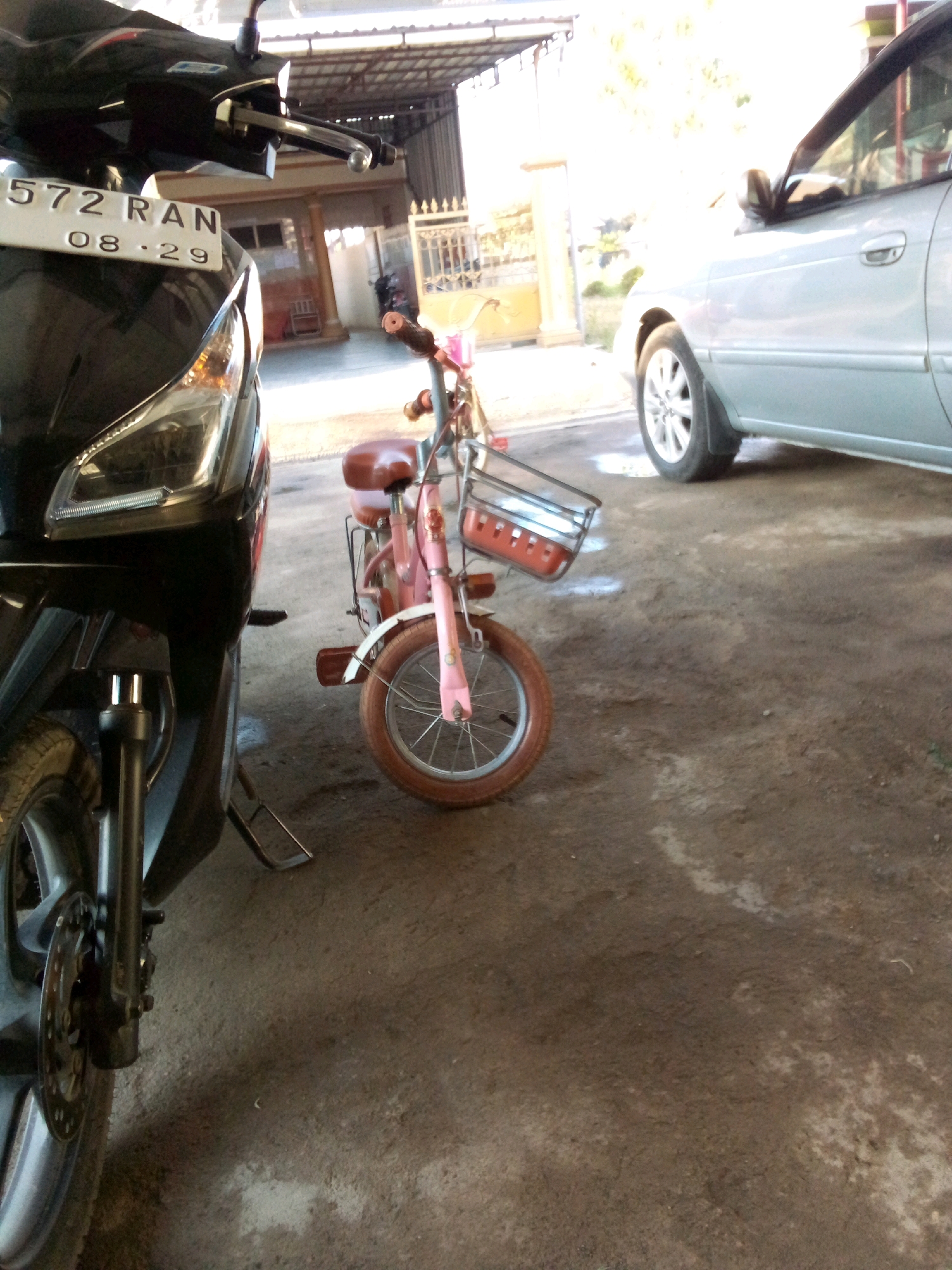 A motorcycle with license plate 572 RAN, dated 08-29, is parked next to a small pink bicycle. The motorcycle is black and the bicycle has a brown seat and a silver basket with red inserts. A silver car is parked behind the bicycle. The scene is outdoors, on a dirt or concrete surface, with a building in the background. The location is Simo, Indonesia.