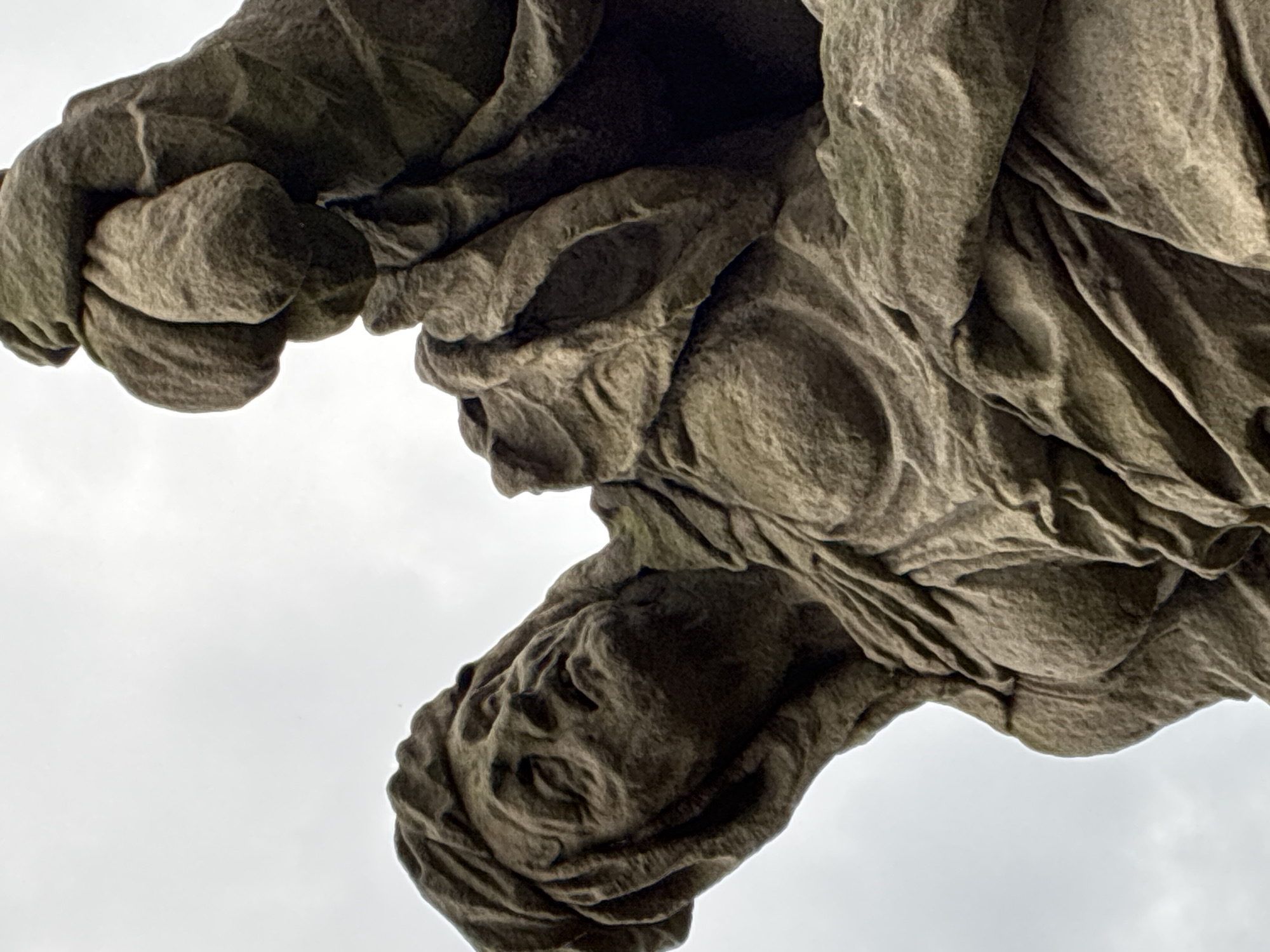 The image shows a close-up, low-angle view of a stone sculpture depicting a weeping female figure. The sculpture is made of weathered stone, with rough textures and visible chisel marks. The figure's head is tilted back, her eyes are closed, and her face is contorted in grief. Her body is draped in flowing robes, which are also rendered in stone with deep folds and creases. One hand is raised towards her face, as if to wipe away tears, while the other hand holds a bundle of fabric or some other object. The background is a pale, overcast sky.
This sculpture is part of the series of statues of allegorical figures that adorn the exterior of the Hospital in Kuks, Czechia, also known as the Kuks Fortress or Kuks Spa. Specifically, this appears to be the sculpture of "Despair" (or "Sorrows") from the series of "Virtues and Vices" created by the sculptor Matthias Braun. The Hospital in Kuks is a Baroque complex located in the Liberec Region of the Czech Republic.
