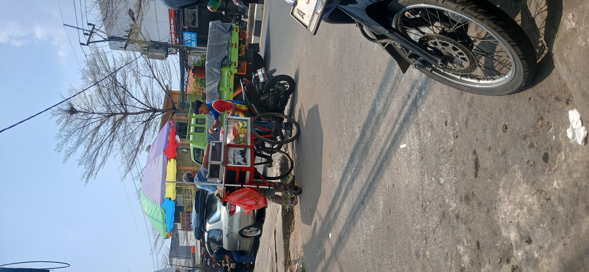 In Baros, Indonesia, on a sunny day, two men are engaged in a transaction at a street food cart. The cart, adorned with a colorful umbrella, is stocked with fruits and vegetables. One man, wearing a blue shirt and gray cap, stands behind the cart, appearing to serve the other. The second man, in a red and blue shirt, is seated on a black motorcycle, reaching towards the cart. A silver car with a roof rack is parked to the left of the cart. In the background, a bright green truck is visible, along with utility poles and buildings. The scene is set on a paved road with shadows cast by the sun, indicating it is likely midday. A second black motorcycle is parked in the foreground.