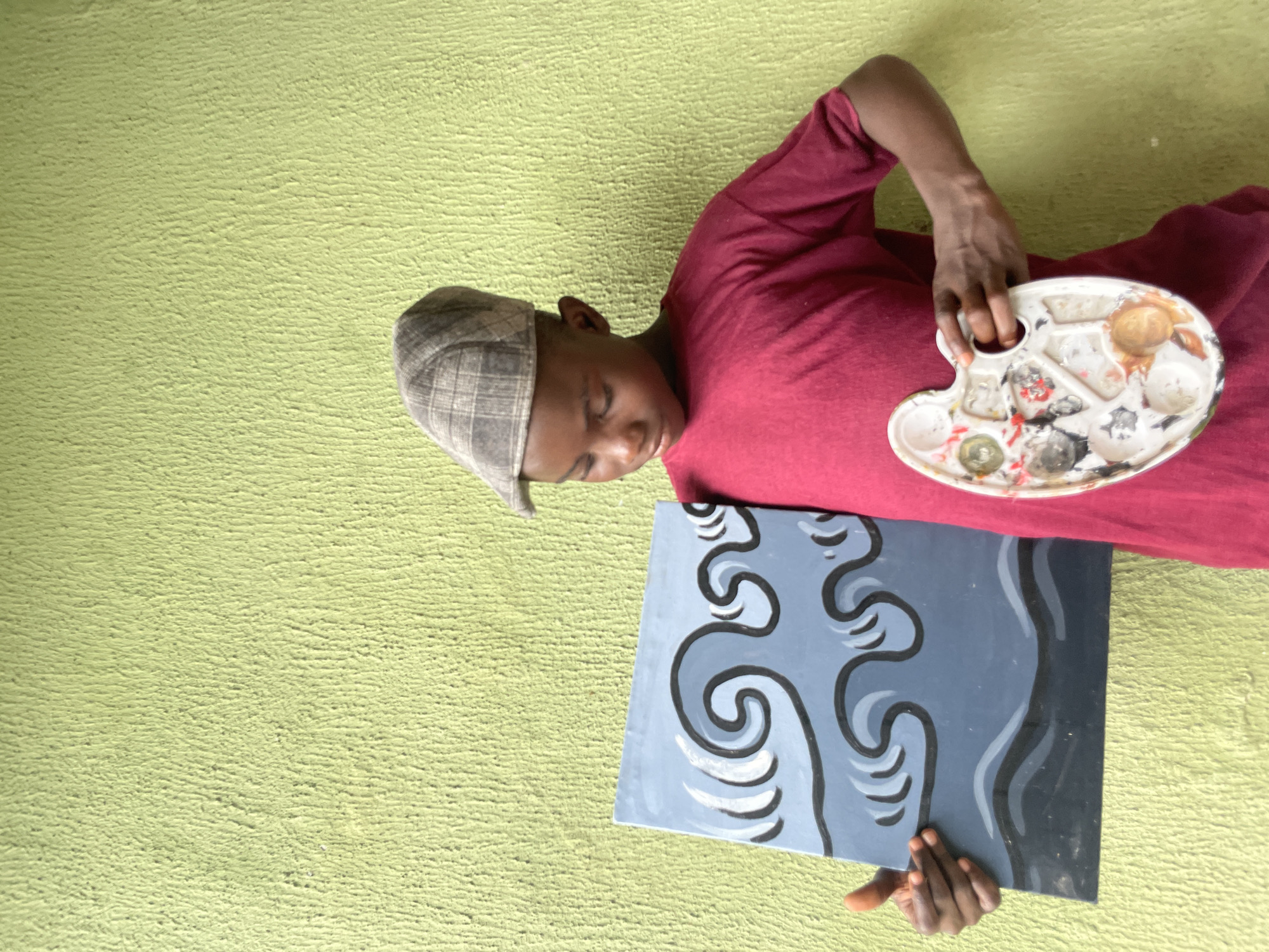 In Ado, Nigeria, a young boy stands against a textured, lime-green wall. He wears a maroon t-shirt and a checkered flat cap. In his left hand, he holds a rectangular painting depicting stylized blue and gray waves outlined in black. His right hand grasps a white artist's palette, which contains various dried paint colors. The boy is looking down at the painting.