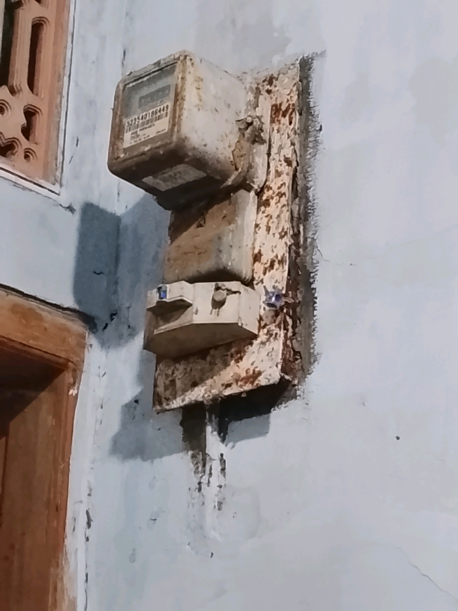 An old, weathered electricity meter and two fused switches are mounted on a light blue wall in Ujungrusi, Indonesia. The meter, boxy and discolored with rust and grime, displays a serial number, "522540186449." The fused switches, also appearing aged and somewhat corroded, are positioned below the meter. The mounting plate behind the electrical components is heavily rusted and peeling. To the left of the meter, a decorative wooden lattice window frame is visible, and a portion of a wooden doorframe is present at the bottom left of the image. The overall scene suggests an outdoor or semi-outdoor environment with evidence of wear and tear.