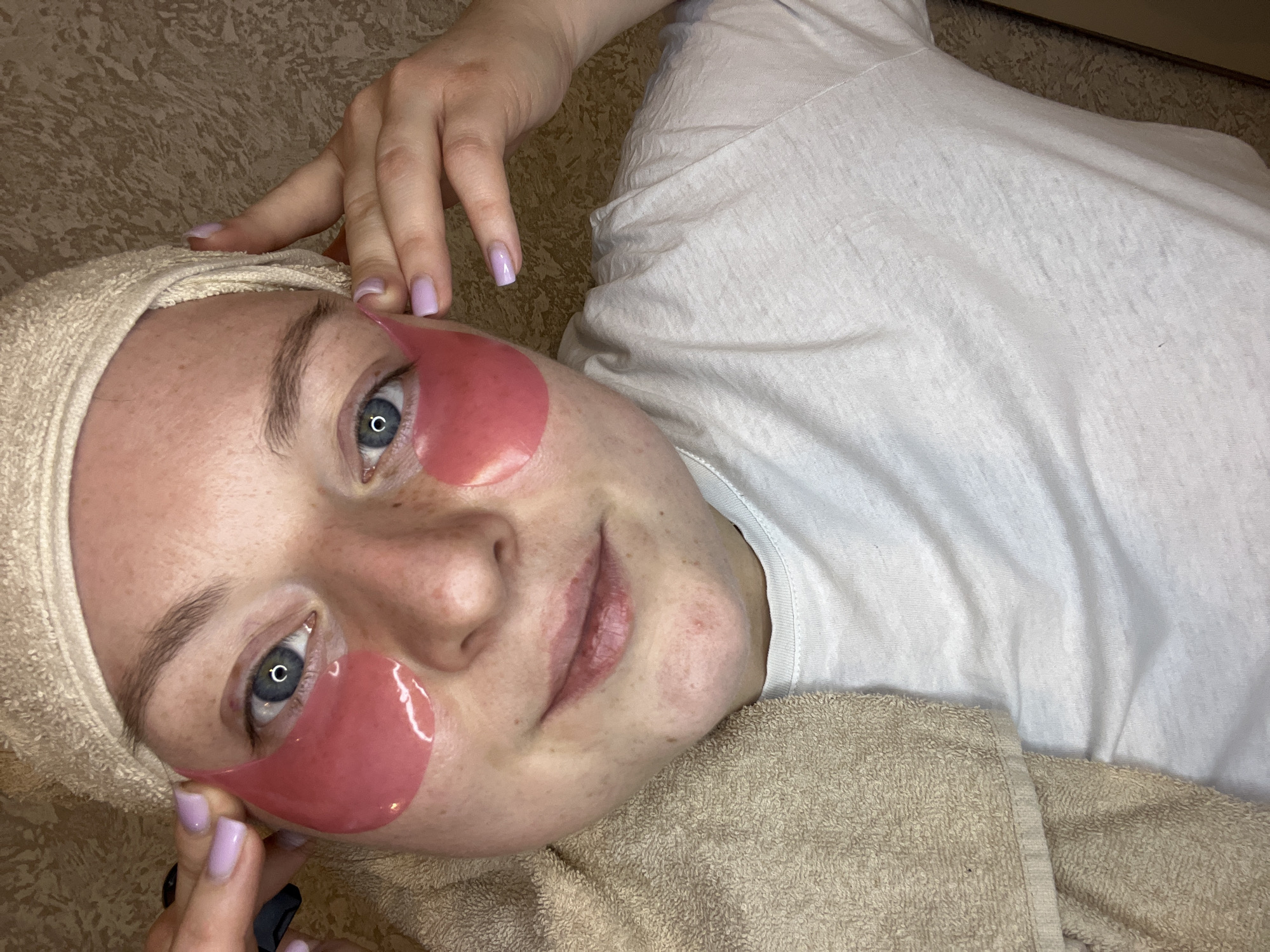 A woman is lying on a surface. She has on two pink eye masks. A light-colored towel is wrapped around her head. She is wearing a white t-shirt. She has light skin with freckles and blue eyes. One of her hands is next to her face and the other is holding the eye mask. The location is not detectable.