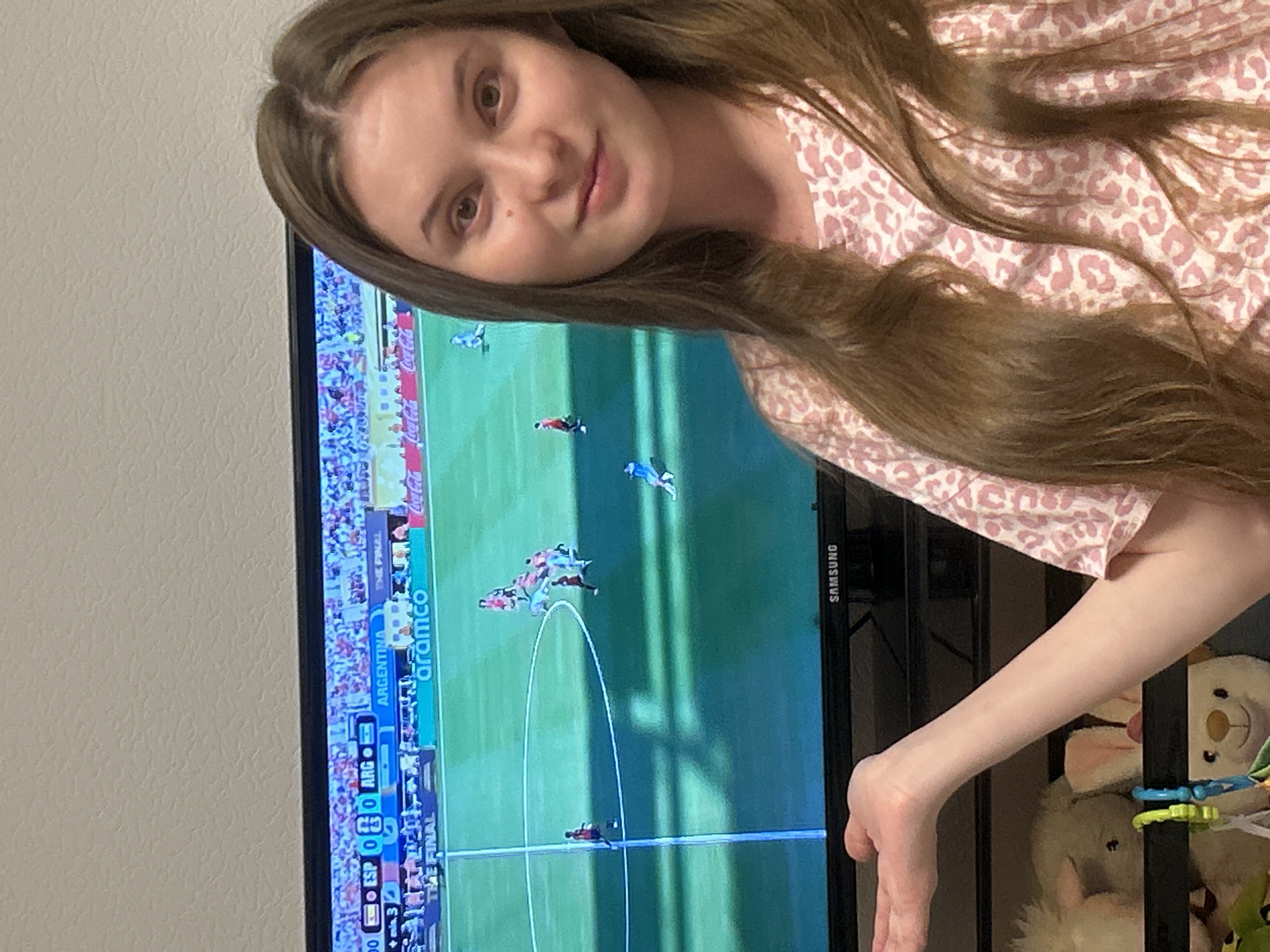 A young woman with long brown hair, wearing a pink and white leopard print shirt, is shown in the foreground of the image. She is looking towards the camera with a slight smile and her left arm is extended with her palm facing outwards, as if gesturing. Behind her, a large television screen displays a soccer match. The score is visible on the screen, showing "ESP 0 - 0 ARG". Several players are on the green field, and advertisements for "aramco" and "Coca-Cola" are visible along the sidelines. The background is a plain, textured wall. The image appears to have been taken indoors. There is no information available about the location in Noviy, Russia.