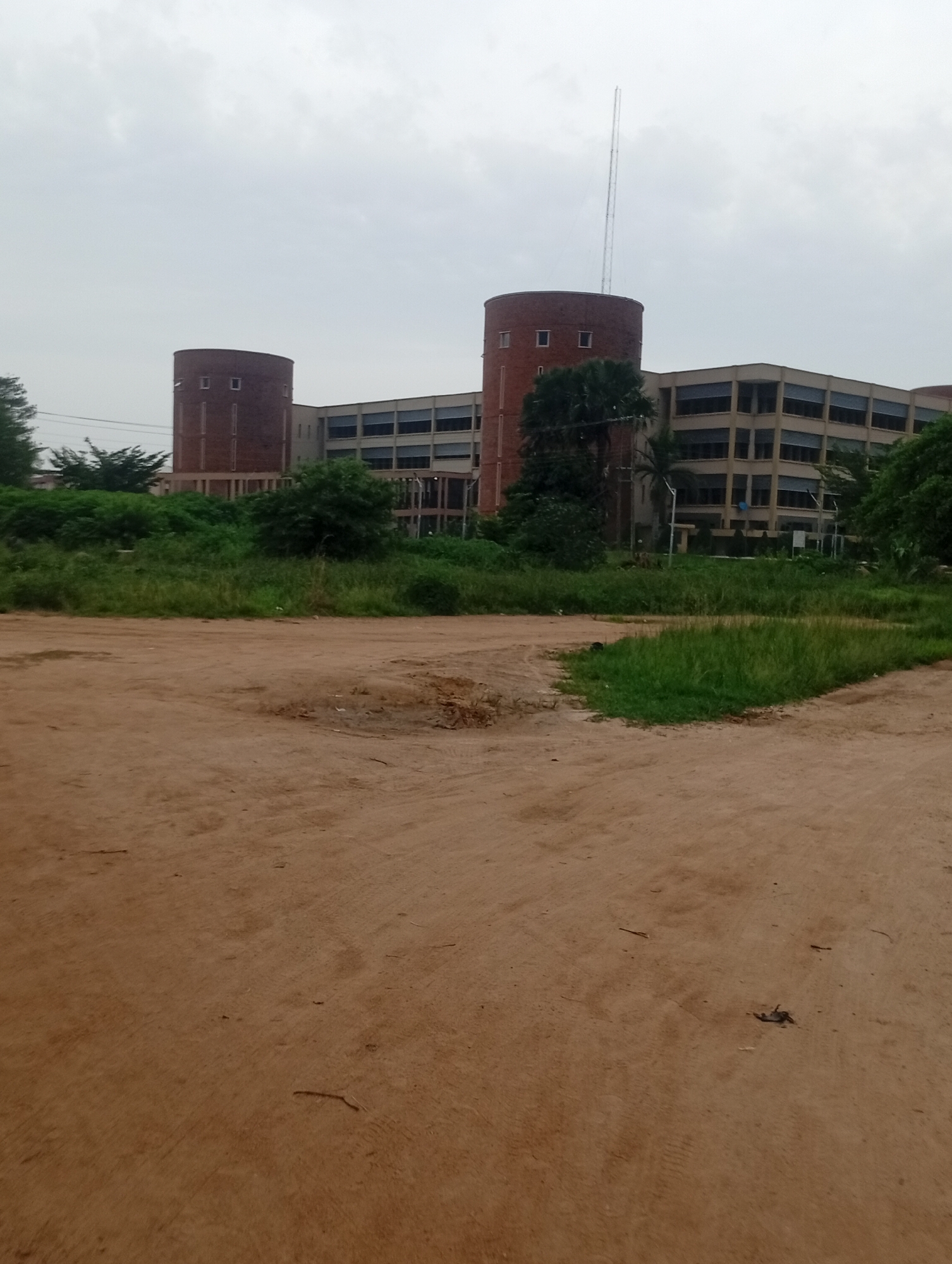 The image captures a wide shot of a modern building complex with a distinctive architectural style, likely situated in Jos, Nigeria. The main structure is a long, multi-story building with a light-colored facade and numerous windows arranged in horizontal bands. Flanking this central building are two cylindrical towers made of dark red brick, each featuring a series of vertical window openings. A tall, slender antenna or mast rises from behind one of the towers.
In the foreground, the scene is dominated by a large expanse of reddish-brown, unpaved ground, possibly a dirt road or a cleared area. The texture suggests it is dry and dusty, with some scattered debris. Beyond the dirt, a strip of green grass and vegetation borders the building complex. This area is uneven and appears somewhat overgrown in places, with various types of trees and bushes visible. The sky is overcast with a pale, diffused light, indicating a cloudy day. The overall impression is one of an institutional or administrative building set within a somewhat undeveloped or natural landscape, typical of certain areas in Jos.