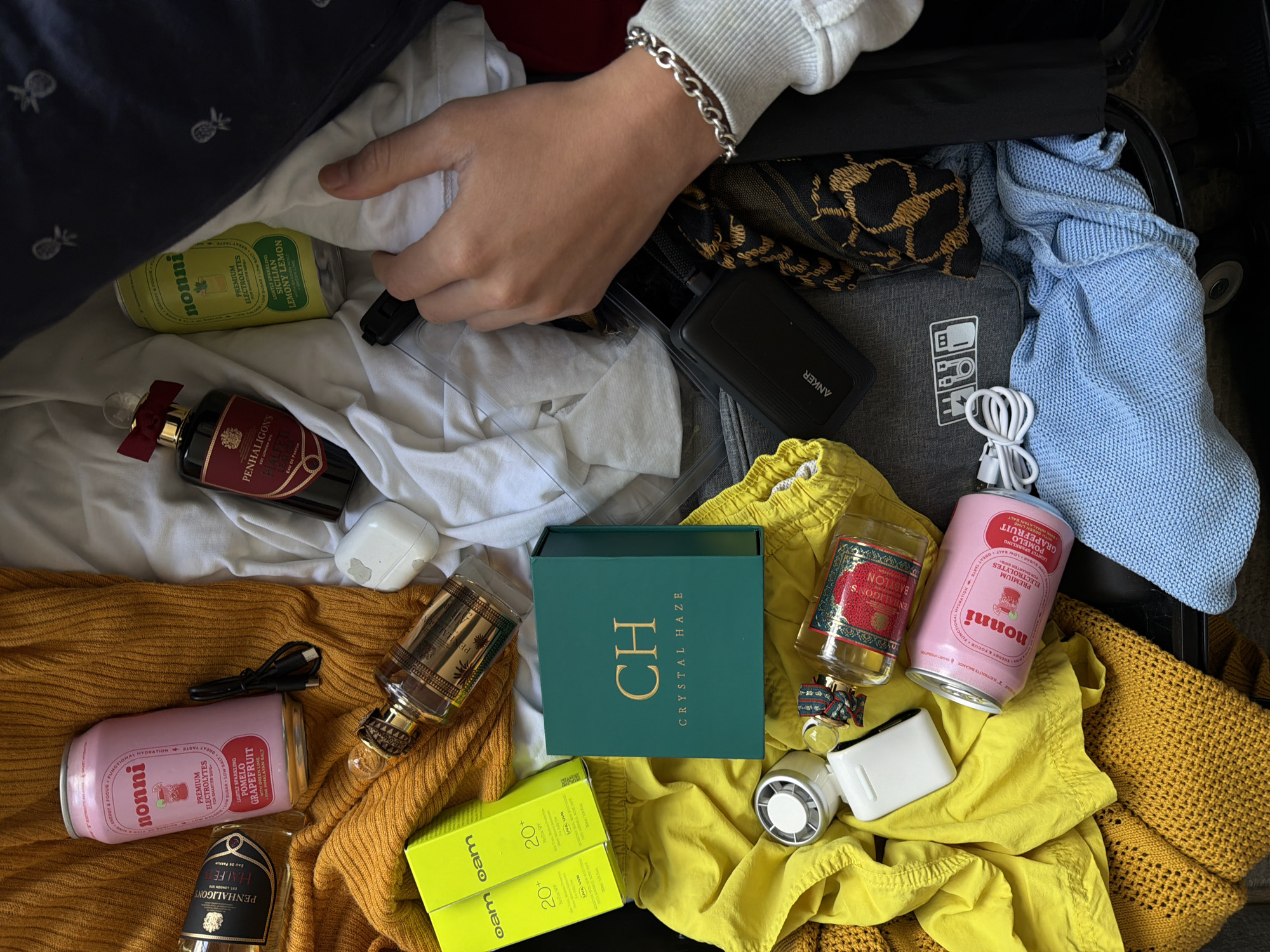 The image depicts a collection of personal items laid out on various textiles, appearing to be packed for travel. The scene is photographed from a top-down perspective. Among the objects are two pink "nonni" electrolyte drinks (one labeled "Pomelo Grapefruit" and the other featuring similar branding), a green box with gold lettering reading "CH CRYSTAL HAZE", two bright yellow boxes labeled "oam oam 20+ SPF", two bottles of Penhaligon's perfume (one labeled "Haute" and another with a dark red label), a white portable fan, a white charging cable, a black Anker power bank, and a navy blue garment with a subtle pineapple pattern. Various clothing items are also visible, including a mustard yellow knitted sweater, a bright yellow pair of shorts, and a light blue knitted blanket. A person's hand, wearing a silver chain bracelet, is visible on the right side of the frame, interacting with the power bank. The overall impression is that of someone meticulously arranging their belongings. The scene does not contain any visual indicators suggesting a specific location in London, United Kingdom, or any urban context.