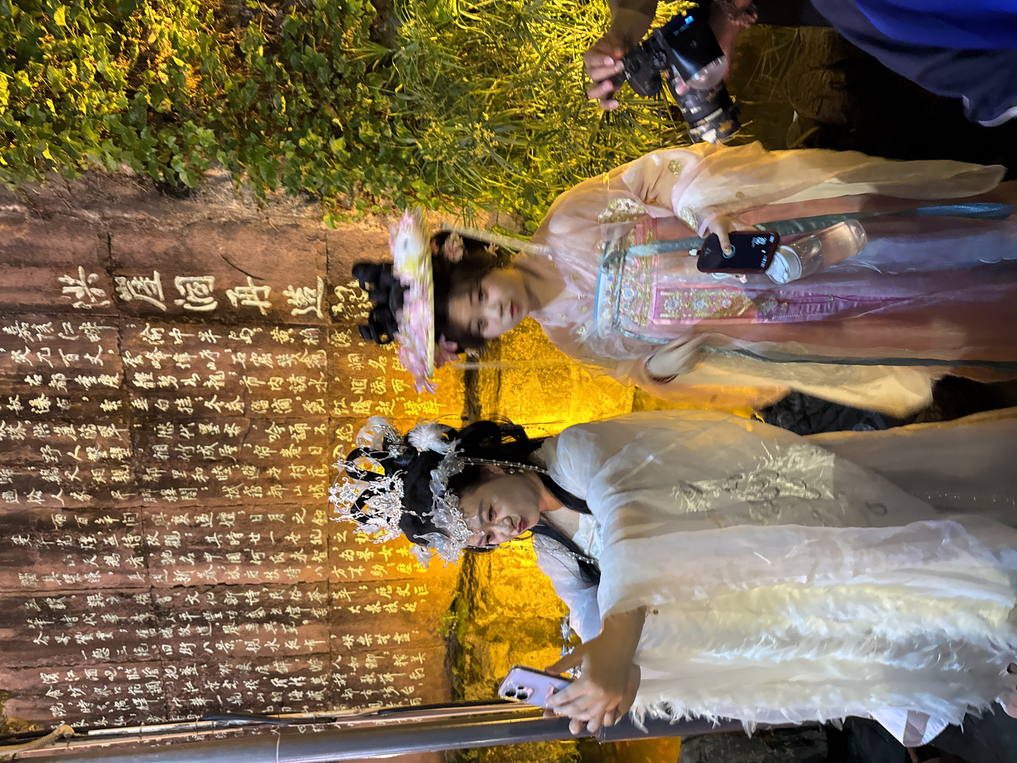 Two women in traditional Hanfu clothing, one in white and one in pink, are posing for photos in front of a stone wall with Chinese characters etched into it. The wall is illuminated with warm light. The woman in white is holding up a smartphone. A photographer is capturing the scene with a DSLR camera. The scene takes place at night, and appears to be in the Yuzhong District, China.