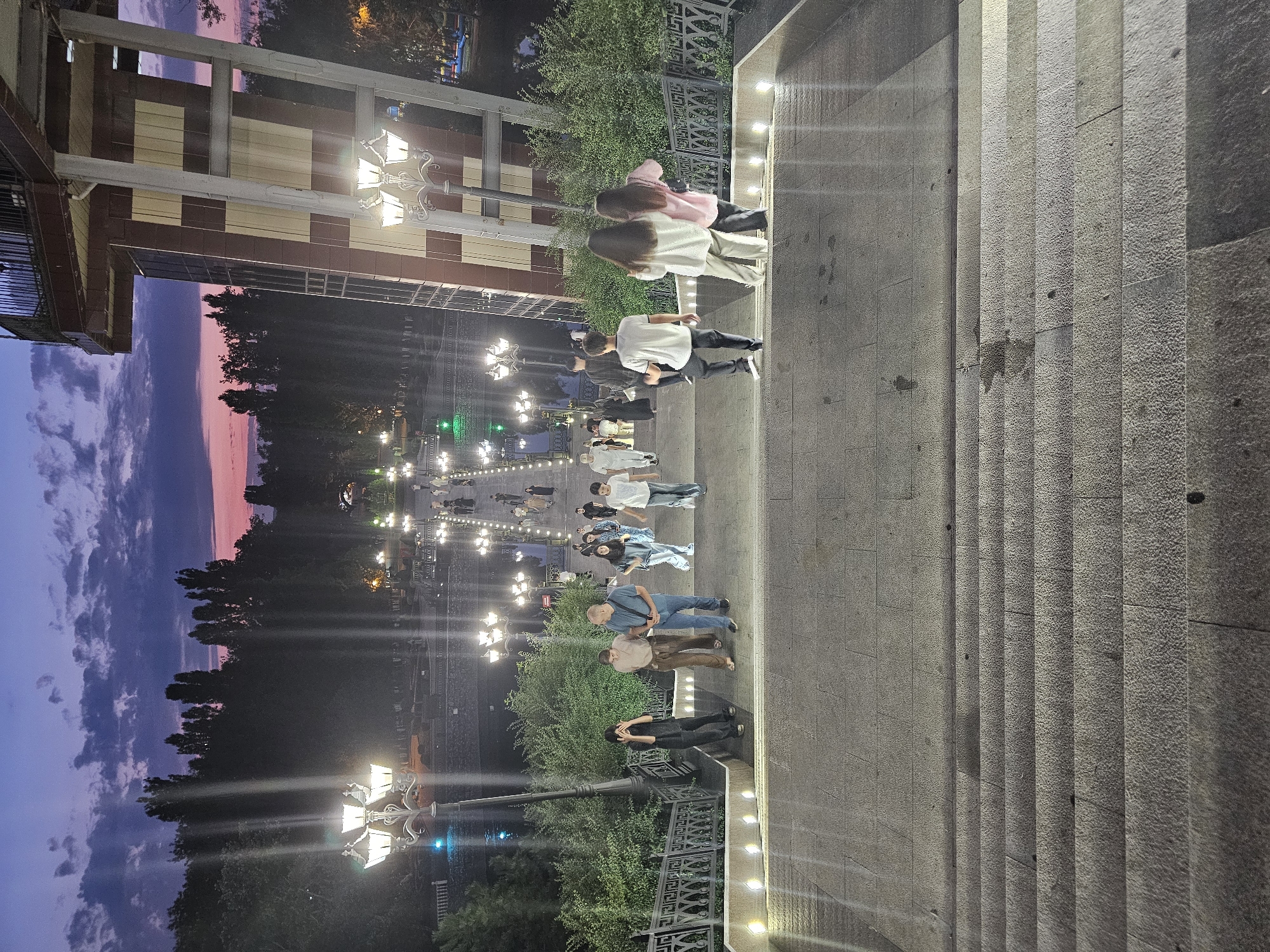 The image captures a twilight scene on a wide, illuminated stone staircase leading down from a modern architectural structure. The sky above is a gradient of deep purple and pink, with scattered clouds. The staircase is bordered by ornate metal railings and illuminated by numerous streetlights, which cast a warm glow on the steps.
In the mid-ground and background, a park or public space is visible, with trees lining a water feature and more decorative lampposts. Several people are present on the staircase and in the park area, walking in various directions.
The architectural structure on the right appears to be a tower or an elevated walkway with a distinctive red and beige patterned facade. Its design incorporates large vertical supports and windows.
The overall ambiance is that of a public space at dusk, with people enjoying the evening. Based on visual cues, this location is identified as the Friendship of Peoples Park in Cherkessk, Russia.