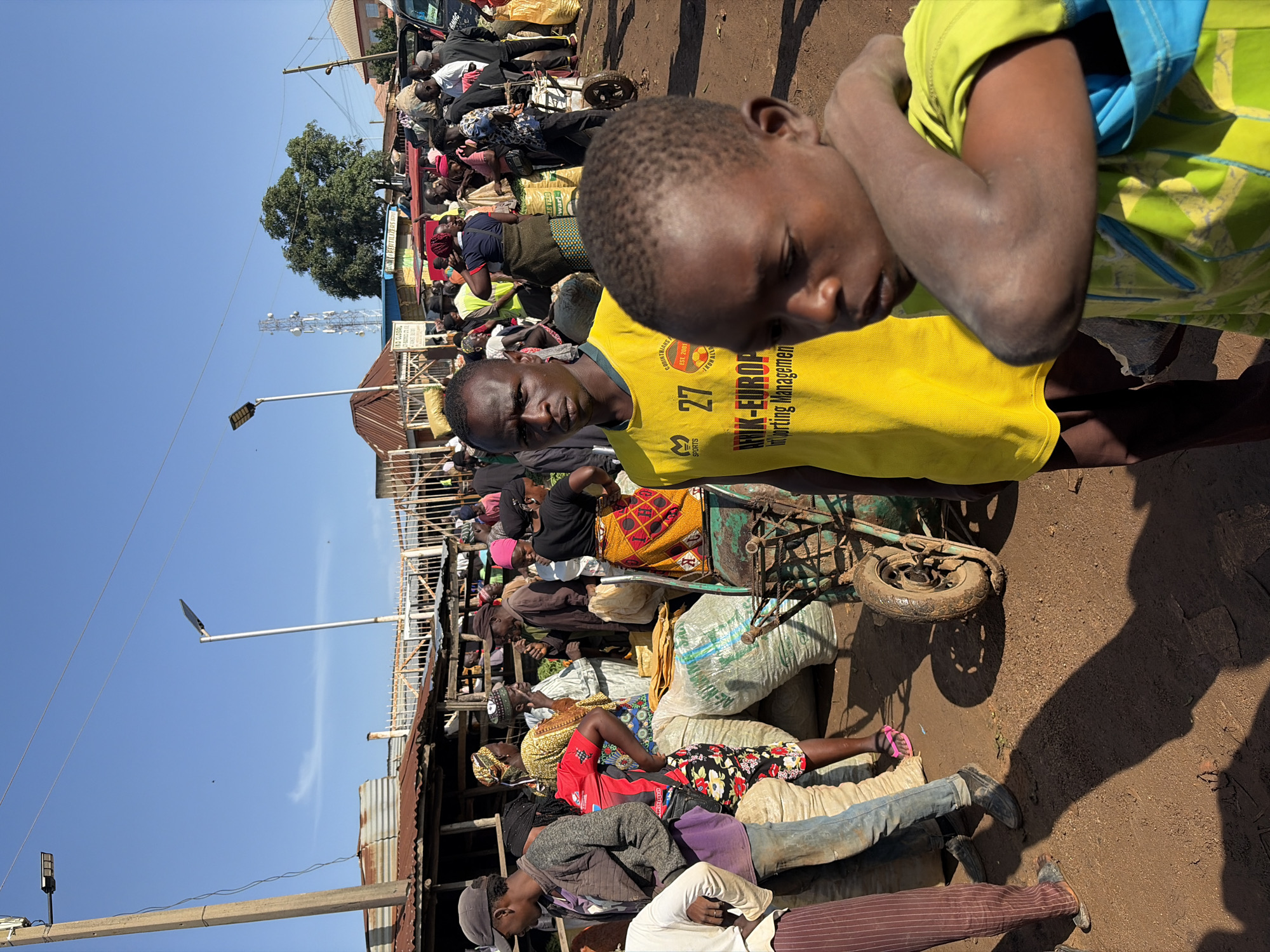 In Gura Top, Nigeria, a bustling marketplace is captured under a clear blue sky. The scene is crowded with people, most of whom are engaged in various market activities. In the foreground, two young boys are the primary focus. The boy on the right, wearing a bright green and blue shirt, is partially obscured, but his arm is raised and resting on his head. To his left stands a young man in a vibrant yellow t-shirt with the number 27 prominently displayed, along with text that reads "AFRIK-EUROPE" and "Sporting Management."
Behind them, the marketplace unfolds with a variety of goods and vendors. Several large sacks, likely filled with produce or grains, are piled up. A man in a red and black shirt is seen interacting with a woman dressed in a colorful, patterned skirt and headwrap. Other individuals are seen browsing, carrying items, or engaged in conversations, creating a dynamic and lively atmosphere. The background reveals more stalls, some with corrugated metal roofs, and hints of residential buildings. Streetlights on tall poles are visible, suggesting infrastructure within the urban setting. The ground is dusty, typical of an unpaved market area. The overall impression is one of a typical day in a busy West African marketplace.