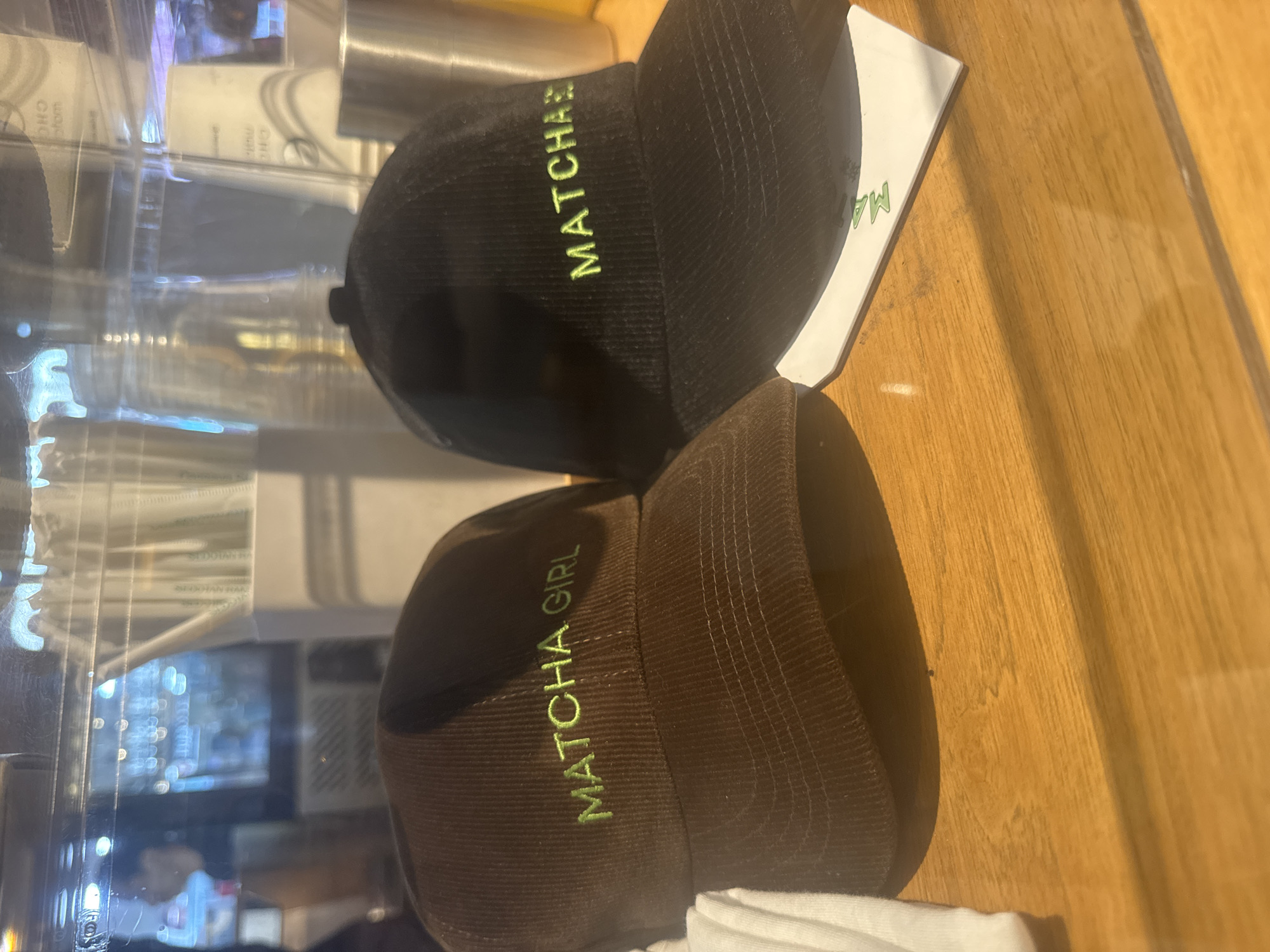 Two baseball caps are displayed on a light brown wooden surface, likely a counter or shelf, in an indoor commercial setting such as a cafe or shop in Kuta Selatan, Indonesia.
The top cap is black with bright green embroidered text that reads "MATCHAS" in capital letters. The bottom cap is dark brown, made of a corduroy-like material, and features the embroidered phrase "MATCHA GIRL" in similar bright green capital letters. Both caps are oriented with their brims pointing towards the right side of the image.
Behind the caps, a clear glass or plastic partition reflects ambient light and indistinct shapes, possibly of containers or other items within the establishment. A white piece of paper or card, bearing partial green text "MA" (likely part of "MATCHA"), is visible underneath the black cap. A fold of white fabric is also partially visible under the brown cap.
The scene depicts merchandise, specifically hats with matcha-themed branding, presented for sale. No people are visible, and the time of day is not explicitly indicated, though the lighting suggests an indoor setting during daylight hours.