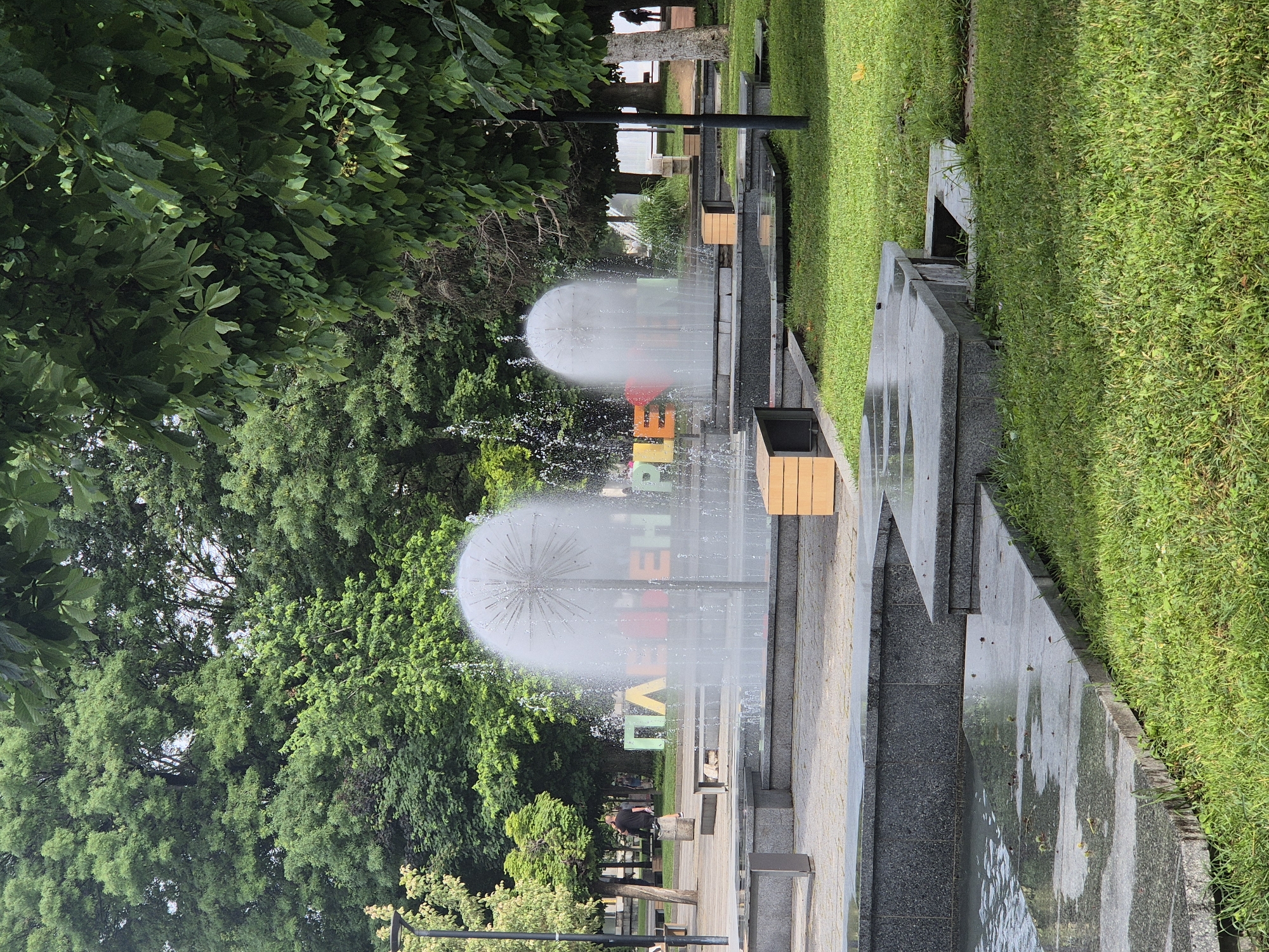 The image displays an upright view of a modern public park or promenade on what appears to be an overcast day. Two large, spherical "dandelion" fountains are prominent in the mid-ground, actively spraying water into the air. Between and behind these fountains, a colorful sign vertically spells out the word "ПЛАНЕТА" (PLANETA, meaning "planet") in capital letters of various hues, including red, orange, yellow, green, and blue. The fountain mist partially obscures some of the letters.
To the left, a dense canopy of lush green trees forms a natural backdrop. To the right, a vibrant green lawn extends, bordered by a multi-tiered concrete or stone structure that incorporates linear water channels and paved walkways. Several people are visible in the background, walking or sitting on benches along the pathways, suggesting a relaxed atmosphere. The pathways feature dark grey paving stones, and there are light wooden elements on what appear to be planters or seating areas. The ground around the water features and fountains appears wet, indicating the constant spray. The lighting is soft and diffused, characteristic of an overcast day, without harsh shadows.