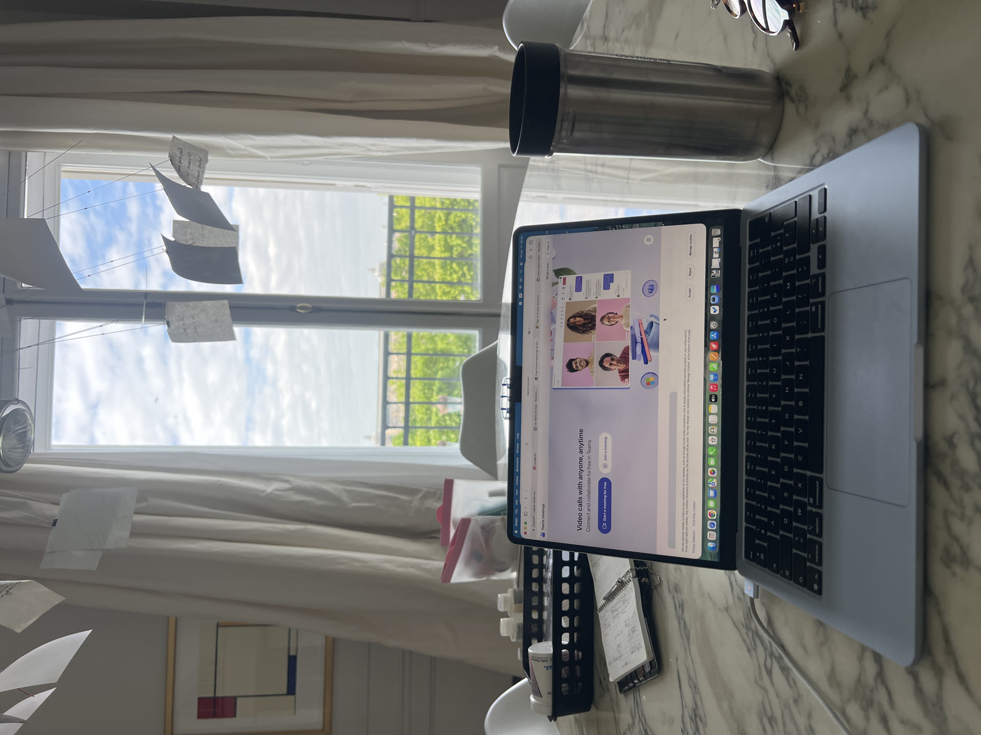 This media file captures a bright, organized workspace by a window, likely in Paris, France.
**Subjects:** The primary objects include a silver MacBook laptop, an iPad displaying the Microsoft Teams website, a silver Nespresso reusable water bottle, eyeglasses, and white curtains. Visible in the background is a window revealing lush green trees and parts of a building exterior under a partly cloudy sky. Inside, several small, handwritten notes hang from strings, creating a mobile-like arrangement. A framed abstract artwork, resembling a Mondrian piece with red, blue, and yellow rectangles, is visible on a wall. Other items on the marble-like desk surface include a black organizer with various small bottles, a spiral-bound notebook, and a charging cable connected to the laptop.
**Activity or event taking place:** The setup suggests a home office or study area. The laptop screen shows the Microsoft Teams website, indicating the user is either preparing for or considering a video conference or collaborative session. The hanging notes might be reminders or part of a creative process.
**Notable details:** The scene is illuminated by natural daylight, indicating it is daytime. The weather outside appears partly cloudy. The general ambiance is functional and tidy.
**Visible text:** On the laptop screen, text from the Teams website is prominent: "Video calls with anyone, anytime", "Connect and collaborate for free in Teams", "Start a meeting for free", and "Join a meeting". Other visible text includes "What's new", "Sign in to your account", "Privacy Statement", and "Third-Party Cookies," along with a cookie consent message. Safari browser tabs show "ChatGPT," "Instagram," and "Les Bains De Spa - Recherch," "Free Online Meetings & Vid." The Nespresso brand name is visible on the water bottle. Handwritten text on the hanging notes and small notebook is illegible.