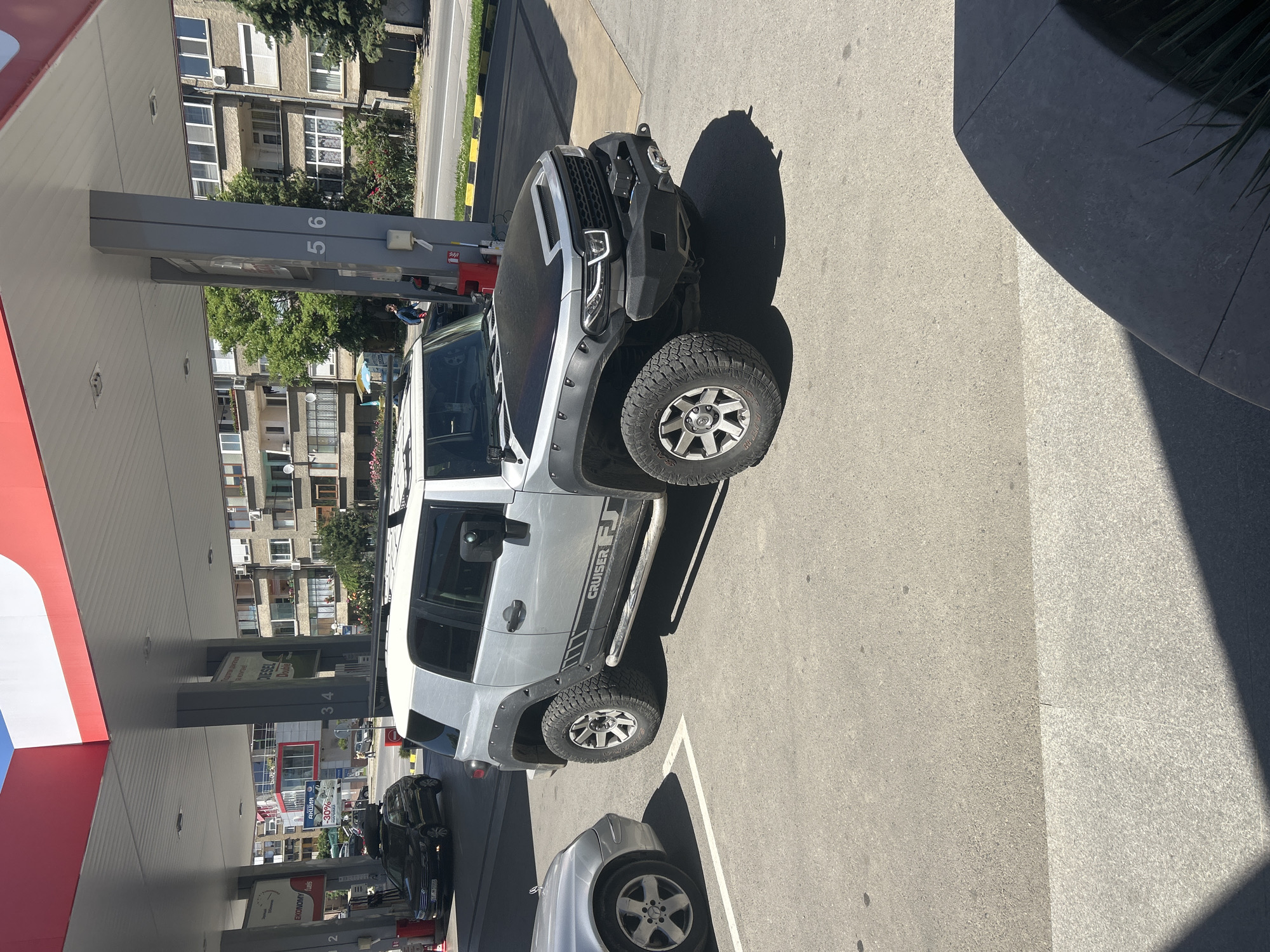 The image shows a silver Toyota FJ Cruiser parked at a gas station. The gas station appears to be in a city with apartment buildings in the background. The car is at a gas pump, with another car parked behind it. The gas station canopy is visible. The city is Kazanlak, Bulgaria. There are numbers on the gas pump marking 5 and 6.