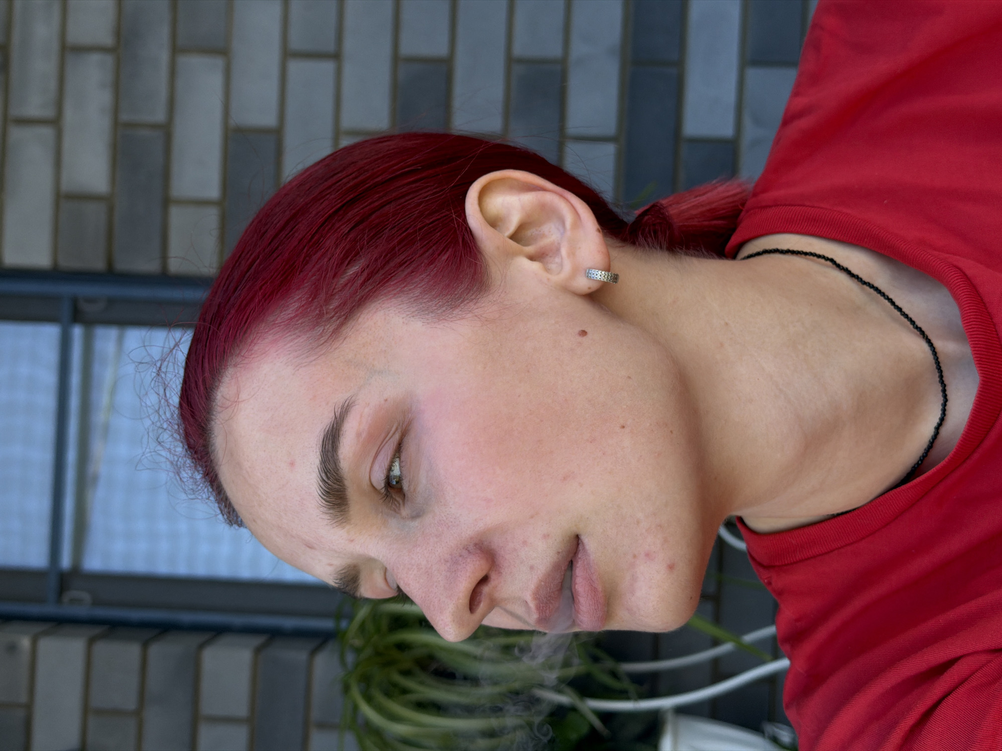 A medium shot captures a young woman with bright red hair, facing left and exhaling smoke, which drifts upwards. She has light skin with some redness and freckles, and her makeup is subtle. She wears a red t-shirt and a black beaded necklace. A small silver hoop earring adorns her left ear. The background consists of a blurred brick wall and a window. The image is taken outdoors, possibly on a balcony or porch, given the visible plant and railing. The setting suggests a residential area. The lighting indicates it is daytime.