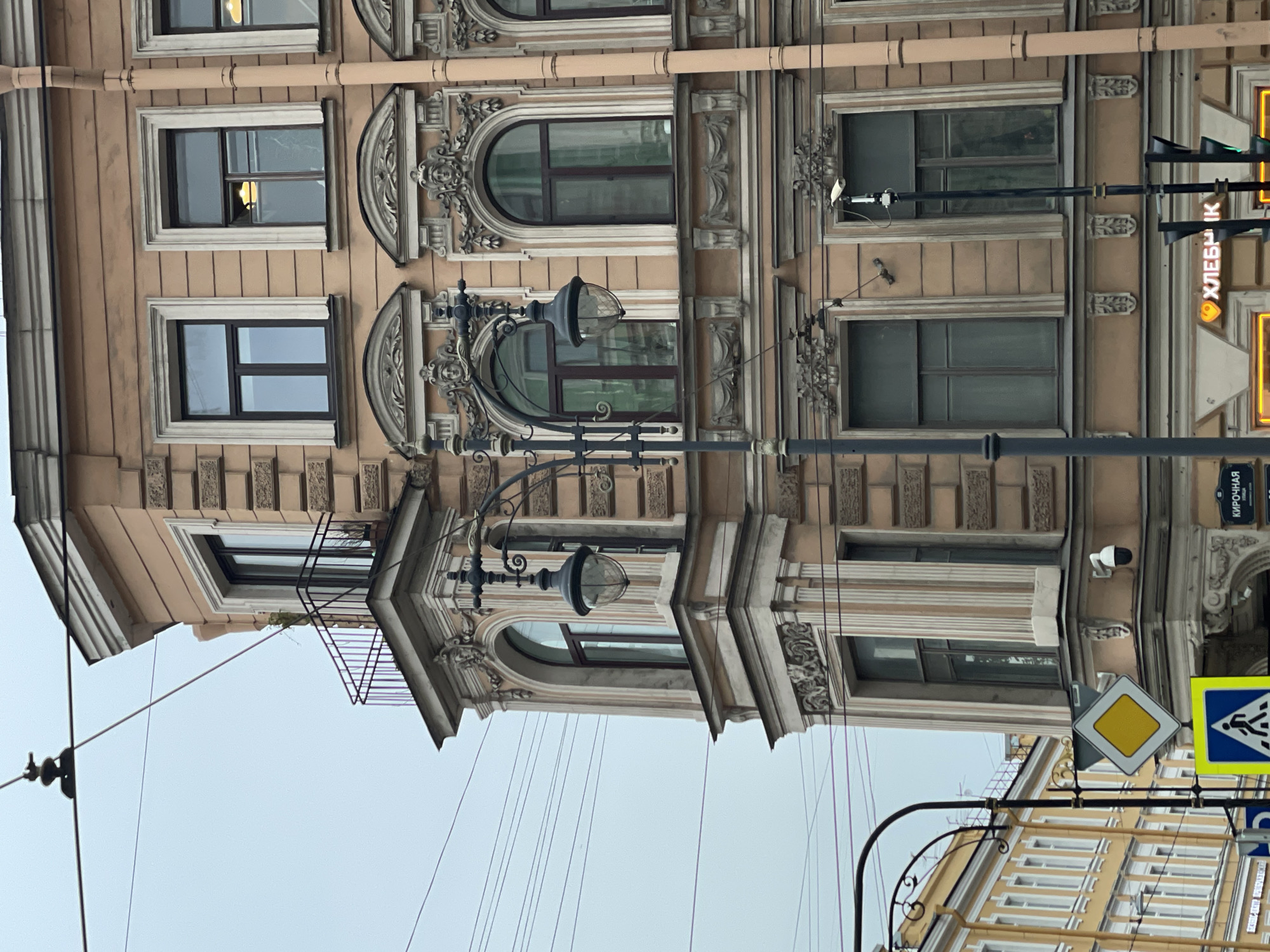 The image captures a building in Saint Petersburg, Russia, showcasing architectural details, street infrastructure, and signage. The building in the foreground exhibits ornate window frames, decorative moldings, and a light tan facade. A street lamp is positioned in front of the building, its lamp head resembling a globe shape, extending from a curved ornate metal support. Overhead utility wires are visible against a grayish sky. In the lower right corner, street signs, including a pedestrian crossing and a yellow diamond-shaped sign, are mounted on a pole. A partially visible building with yellow and white window frames is in the background. The text "ХЛЕБНИК" (meaning "bread maker" or "baker" in Russian) is visible on a vertical sign in yellow and white, indicating a local business. A security camera is affixed to the building in the background. The overall scene depicts an urban environment with a blend of historical architecture and modern street fixtures.