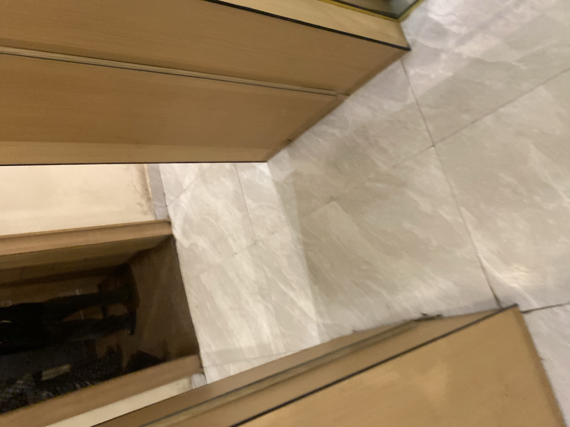 In the city of Sabon Gari West, Nigeria, the image depicts the interior of a residence. The foreground shows a polished, light-colored tiled floor. To the left, a portion of a wooden doorframe is visible, with its surface reflecting a dark, indistinct image of a person's legs and feet. To the right, a tall, light-brown wooden wardrobe stands, with its doors closed. Further in the background, another wooden doorframe leads into a darker room where the reflected image of the person is clearer, showing them wearing dark trousers and shoes. The walls appear to be painted a pale yellow color. The overall impression is one of a dimly lit hallway or interior space within a home.