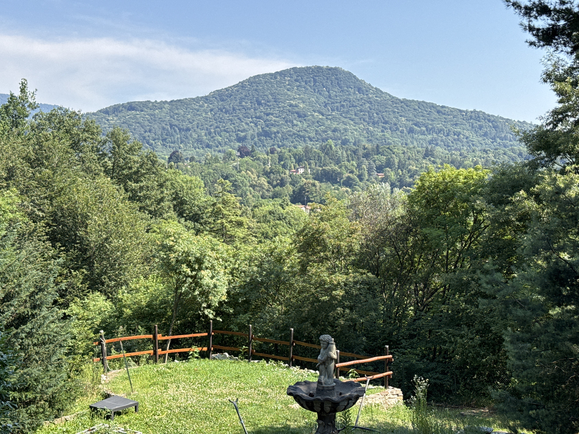 The image shows a mountainous landscape in Cunardo, Italy, on a clear day. In the foreground, there is a lush green lawn with a stone fountain featuring a statue of a child holding a stalk. To the left of the fountain, a dark, rectangular object, possibly a low table or bench, sits on the grass. A wooden fence with horizontal rails runs across the middle ground of the image, separating the lawn from the forested slopes behind it. The middle ground is filled with dense, green trees, with a few scattered houses visible amongst them. The background is dominated by a large, tree-covered mountain that rises majestically against the bright blue sky. The overall scene depicts a serene, natural environment.