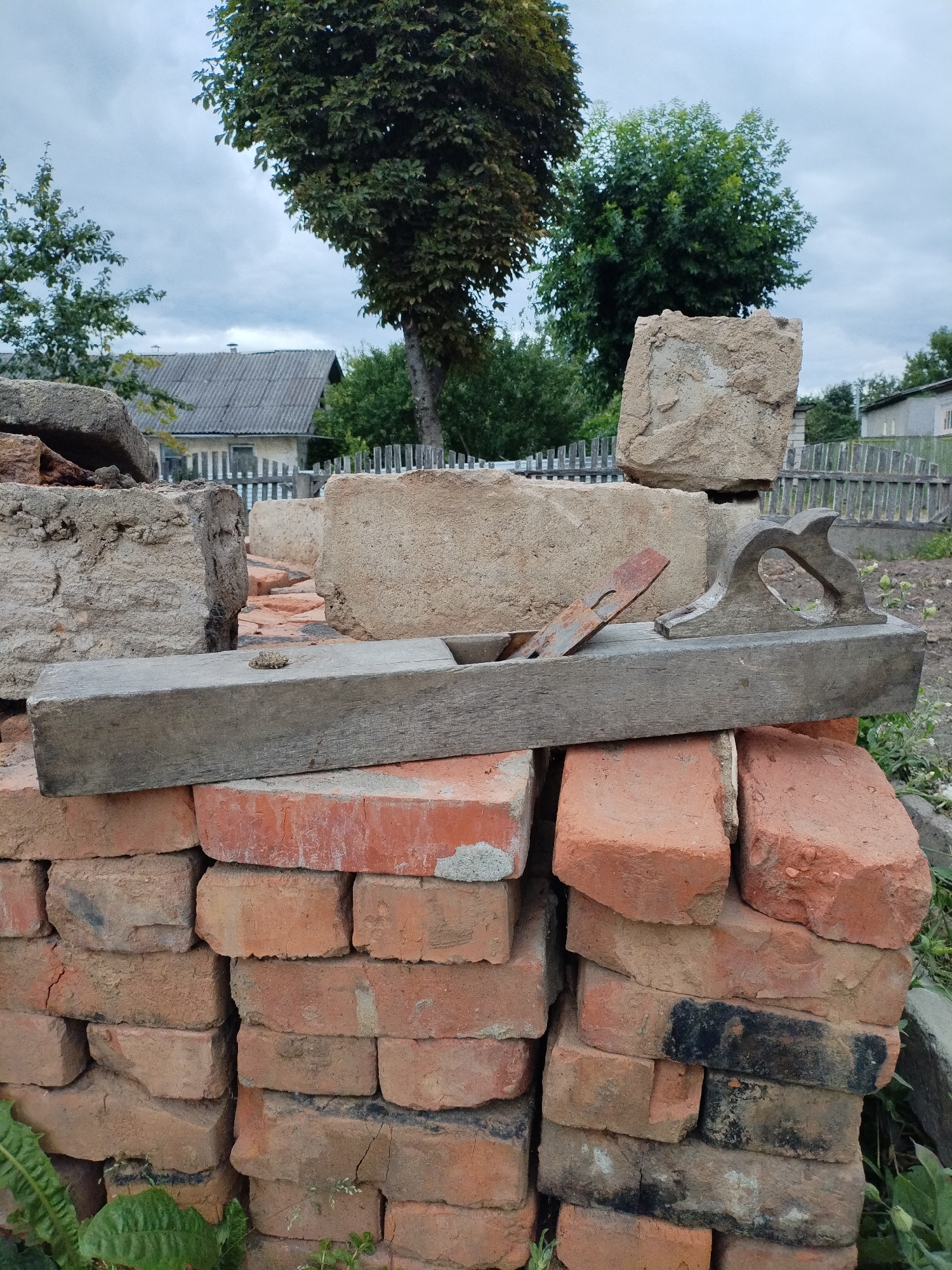 The image depicts an outdoor scene, possibly a construction site or a yard, with various building materials and vegetation. In the foreground, a stack of red bricks is visible, supporting a weathered wooden hand plane. The hand plane has a rusty metal blade. Behind the bricks and plane, larger concrete blocks and stones are arranged, some of which appear to be weathered and broken.
In the background, a residential building with a grey, corrugated roof and white trim is partially visible, along with a wooden fence. Trees with green foliage are scattered throughout the scene, suggesting a rural or suburban setting. The sky is overcast with grey clouds. The image is taken from a slightly elevated perspective, looking towards the background. There are no people or animals present in the frame. The location is identified as Bialynichy, Belarus.