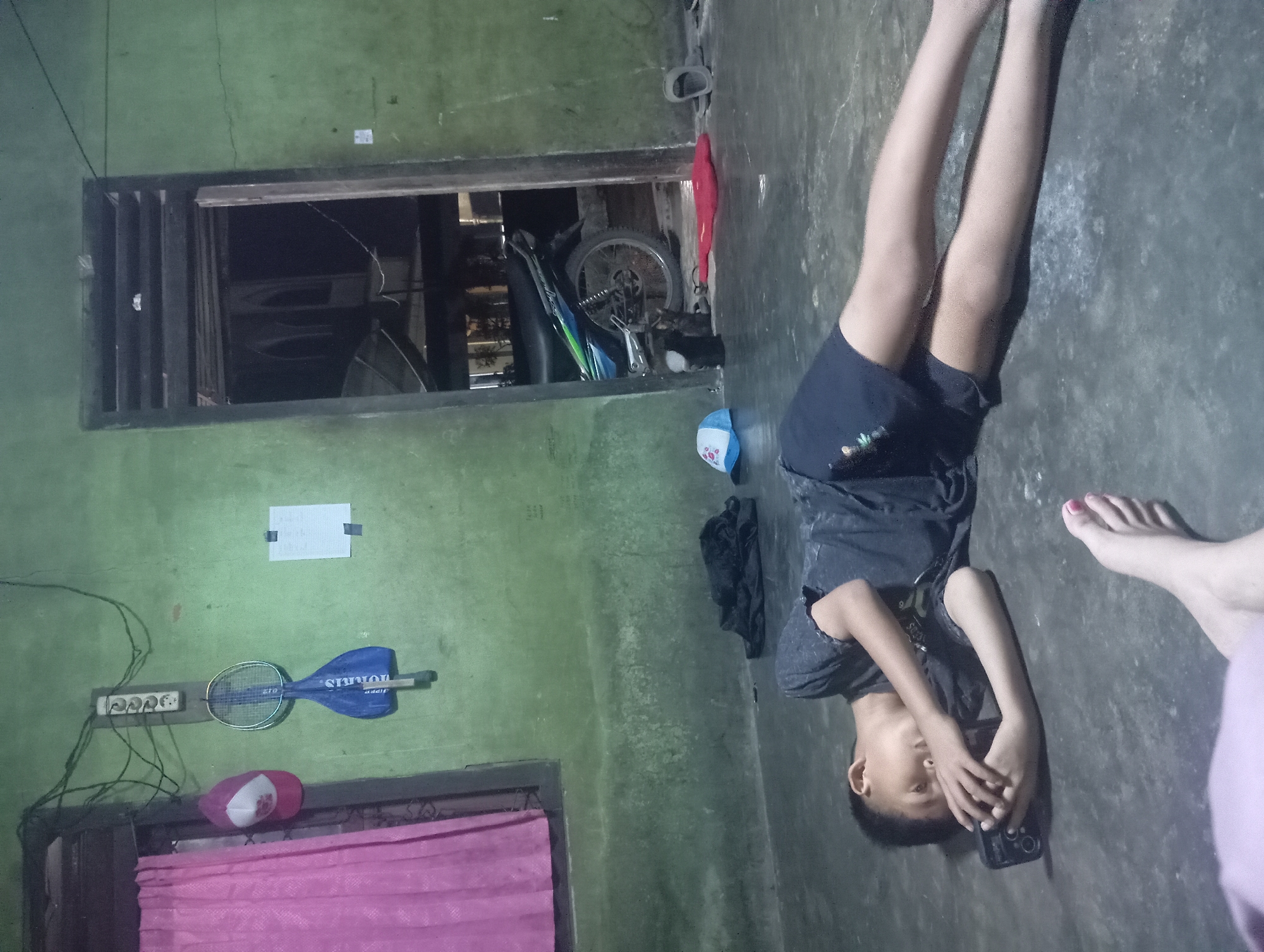 A young boy lies on a concrete floor, propped up on his elbows, looking at a smartphone he holds in his hands. He is wearing a dark t-shirt and dark shorts. His body is extended to the right. In the foreground, the feet of another person are visible, with one foot having painted toenails. The room has green walls and a dark concrete floor. To the left, a pink curtain hangs over a window. On the wall above the window, a power strip with multiple outlets is mounted, with wires extending from it. A badminton racket in a blue bag hangs from the wall next to a pink and white baseball cap. A white paper with some writing is taped to the wall. In the background, an open doorway reveals a dark interior with a motorcycle parked inside. A red object and a blue cap are on the floor near the doorway. The overall scene suggests a casual, indoor environment. The location is identified as Rejosari, Indonesia.
