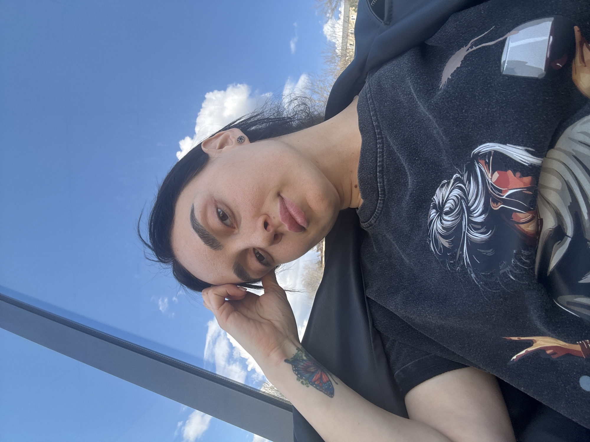 A woman is pictured from a low angle in Tbilisi, Georgia, on what appears to be a sunny day. She is wearing a dark-colored t-shirt with a graphic design, and a black jacket is draped over her shoulders. She has dark hair and noticeable eyebrows. Her right hand is raised to her head. A butterfly tattoo is visible on her left forearm. The background shows a blue sky with some white clouds, partially framed by a reflective surface, possibly glass, of a building or structure. The scene appears to be outdoors.