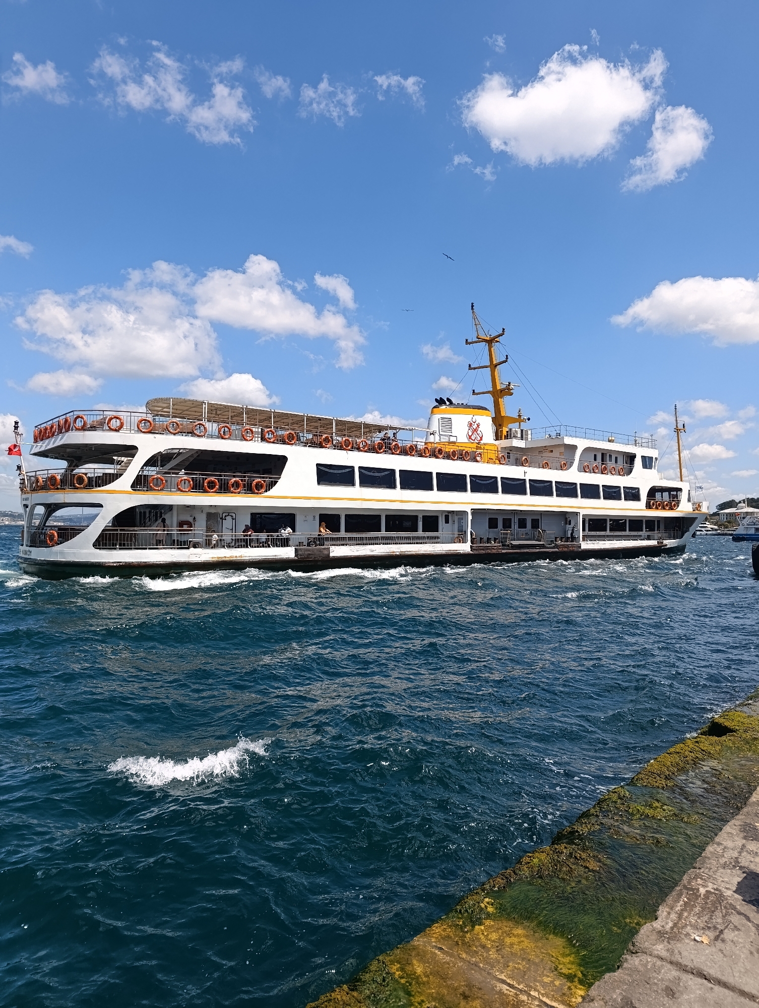 A white ferryboat with yellow accents is cruising on choppy, deep blue water under a bright blue sky with scattered white clouds. The ferry has multiple decks, with life preservers visible along the railings of the upper levels. Several people are visible on the decks, some standing and others seated. A tall yellow mast with antennas and navigational lights stands at the stern of the boat. The water is disturbed by the ferry's movement, creating white wakes. In the foreground, a textured stone quay or pier, covered in green algae and moss, lines the right side of the image. In the distance, along the horizon, hints of buildings and another boat are visible. The overall impression is a sunny day on the water in Istanbul, likely the Bosphorus Strait.