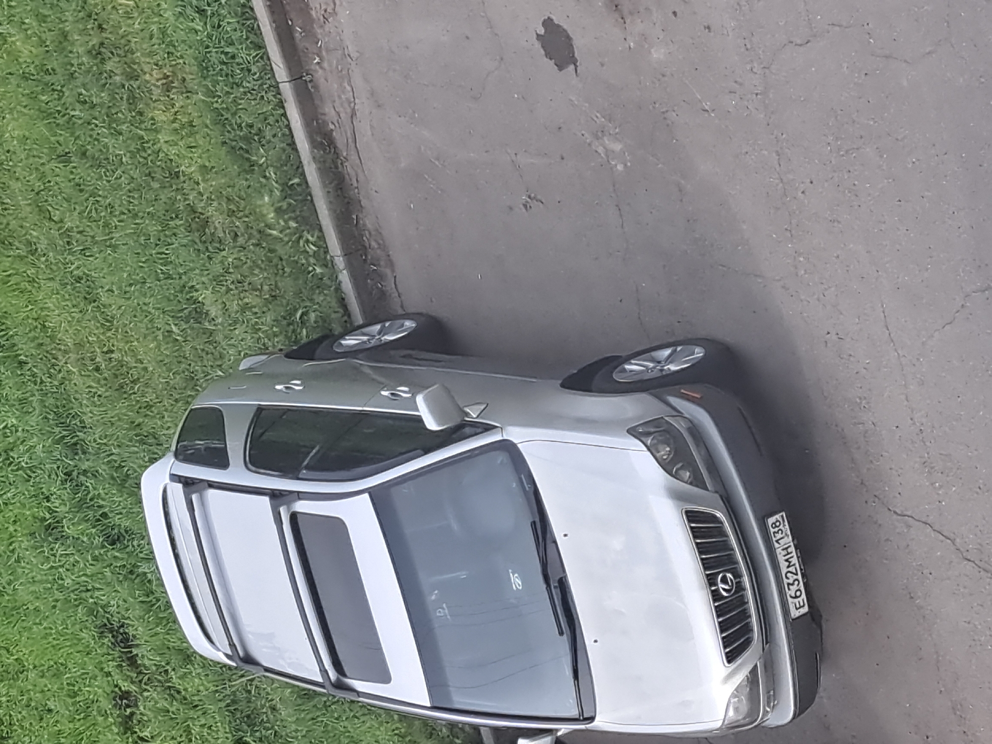 A silver Lexus RX SUV is parked on an asphalt surface next to a strip of green grass. The car has a sunroof and roof racks. The license plate reads "E632MH 138". The background consists of a curb and more grass. The image appears to have been taken from an elevated position, looking down at the car. The scene is consistent with a residential or public parking area in Irkutsk, Russia.