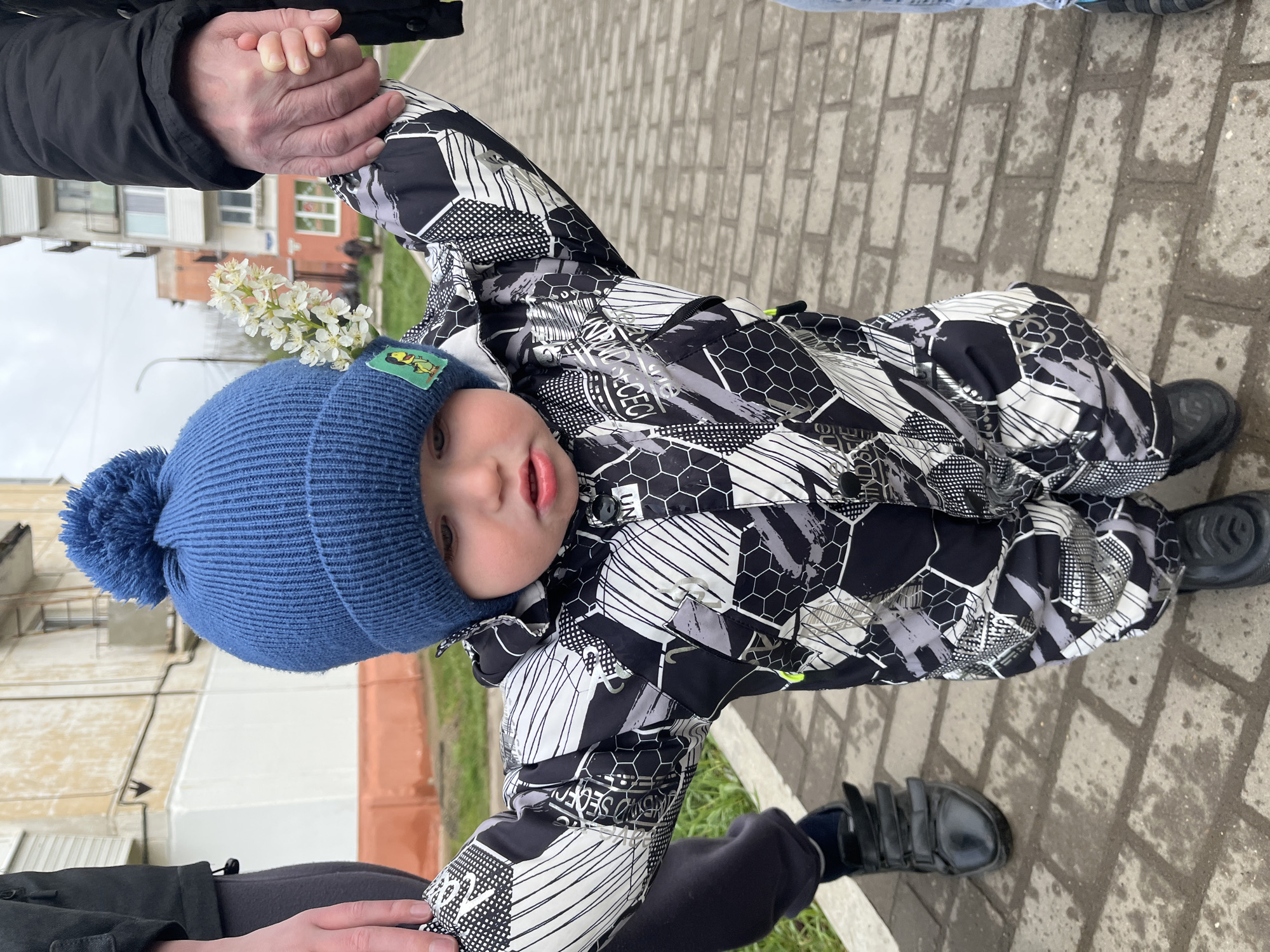 A child in a blue knit hat and patterned black-and-white snowsuit is seen from a slightly elevated angle, standing on a brick sidewalk in Chaikovsky, Russia. The child is being held by the hands of two adults, one visible on either side of the child. The child has a sprig of small white flowers tucked into the hat. Buildings are visible in the background, along with some grass. The sidewalk is wet and has a pattern of rectangular bricks.