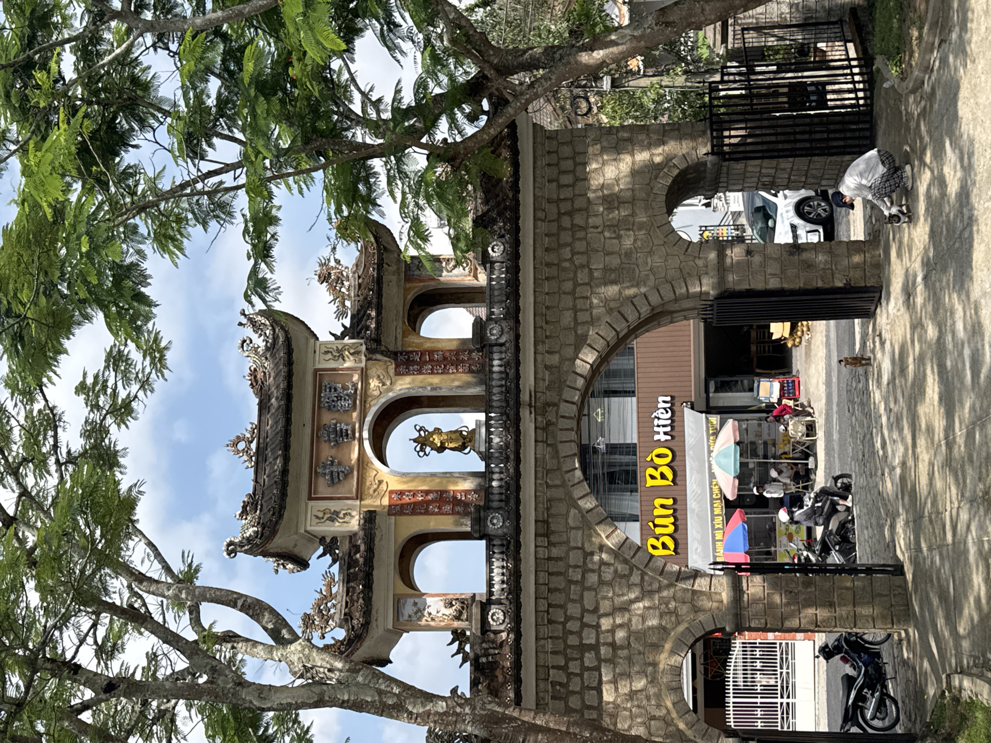 The image depicts an ornately decorated gateway leading to a business in Da Lat, Vietnam. The gateway, constructed of stone, features multiple arches, with a traditional Vietnamese-style roof embellished with intricate carvings and figurines above the central opening. A gold-colored statue is visible through the central arch.
To the right of the gateway, visible through the arch, is a storefront identified by a sign that reads "Bún Bò Hiền" in large yellow Vietnamese script. An additional line of text reads "BÁNH MÌ XÍU MẠI CHÉN MÁ DỪA TƯƠI". Tables and chairs are set up near the storefront, with people seated at some of the tables. Several parked motorcycles are nearby.
To the right of the gateway, a person is crouched down. The person is interacting with a cat. A white vehicle is behind them.
Trees with lush foliage frame the left side of the image, partially obscuring the sky and structure.