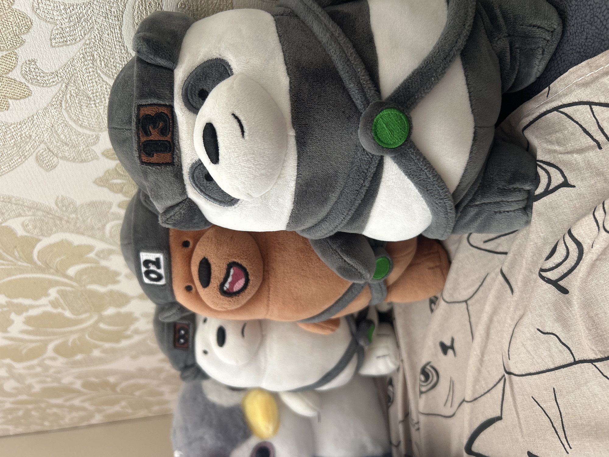 The image shows a row of four plush toys lined up on a bed. From left to right, the first toy appears to be a grey penguin with a yellow beak and pink eyes. Next to it is a white bear with black eyes, wearing a grey cap with "02" in white on a black square. The third toy is a brown bear, also wearing a grey cap with "02" on it, and has a wide, open-mouthed smile. The fourth and closest toy is a panda bear wearing a grey cap with "13" in black on a brown square. This panda also has a grey scarf-like accessory with a green circular embellishment. The toys are positioned on a patterned bedspread with black line drawings of cats. The background is a textured wallpaper with a light floral pattern. The image does not contain any elements that would place it in the city of Khyrdalan, Azerbaijan.