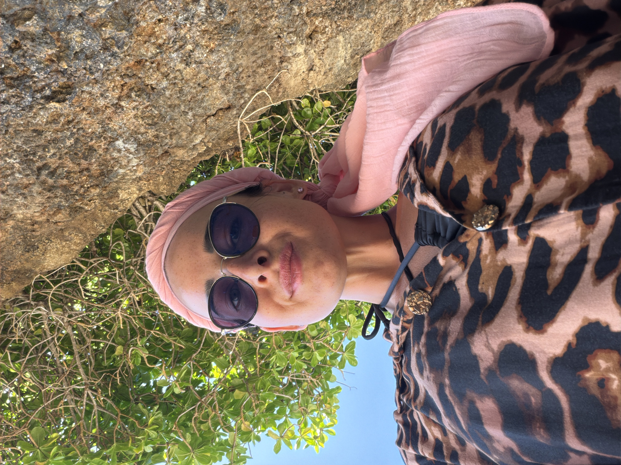 A person is pictured in what appears to be an outdoor setting. The subject is wearing a pink headscarf, sunglasses, and a leopard-print top with decorative buttons. Above and to the left of the person is a large rock or boulder. In the lower portion of the image, there is green foliage and a glimpse of a clear, blue sky. This scene appears to have been taken in the city of Kutuh, Indonesia.