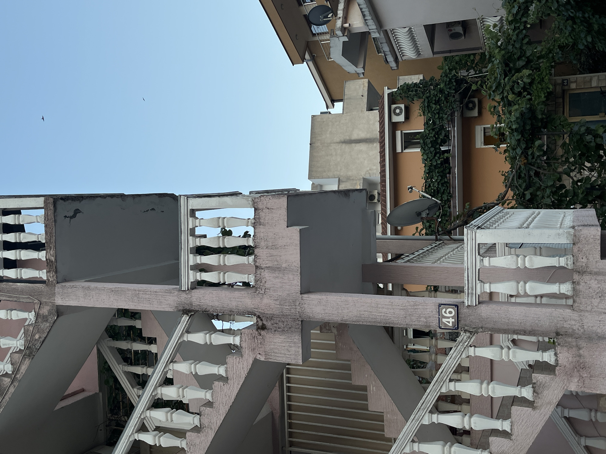 This image shows an outdoor view of a building in Budva, Montenegro, featuring a prominent pink-tinted concrete exterior staircase with white balusters. The staircase winds upwards against a textured concrete column. A small, dark-colored plaque with the number "46" is affixed to the column. In the background, several other buildings are visible, some with orange facades, satellite dishes, and air conditioning units. Lush green foliage, likely vines, covers parts of the buildings. The sky above is clear and light blue, with two birds visible in flight. The scene depicts a residential area with elements of architectural design and natural overgrowth.