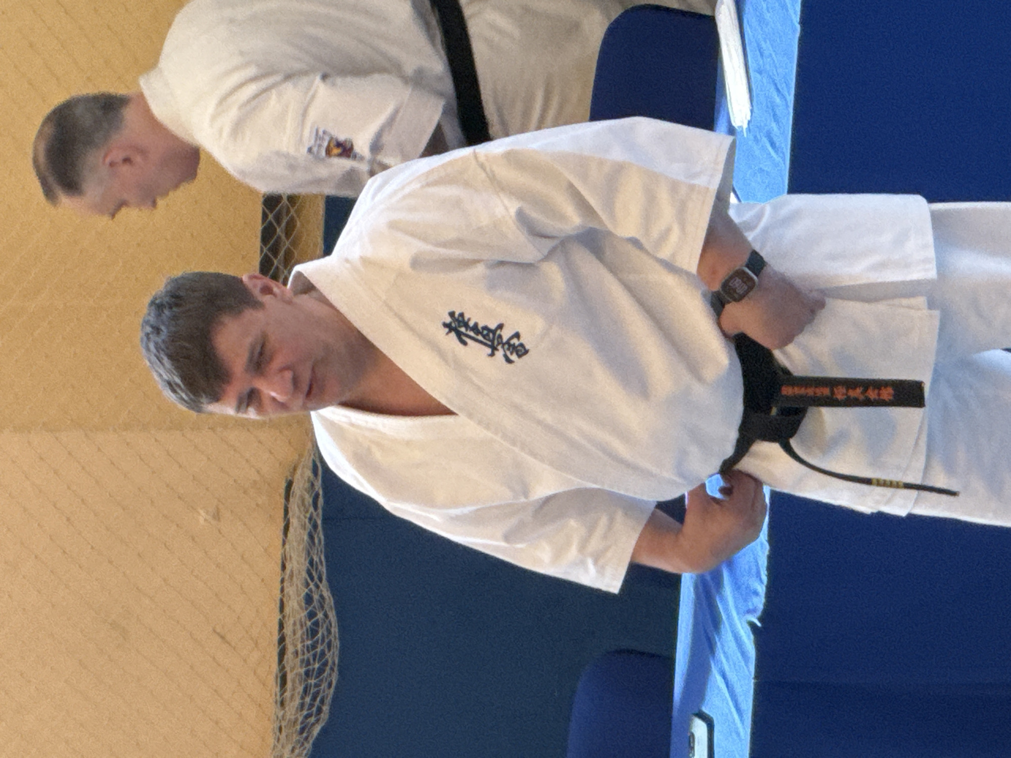 Two men are in a room with a blue background. The man in the foreground is wearing a white karate uniform with a black belt and a watch on his left wrist. He is holding the belt in his hands. A second man is bowing in the background. The location is Yekaterinburg, Russia.