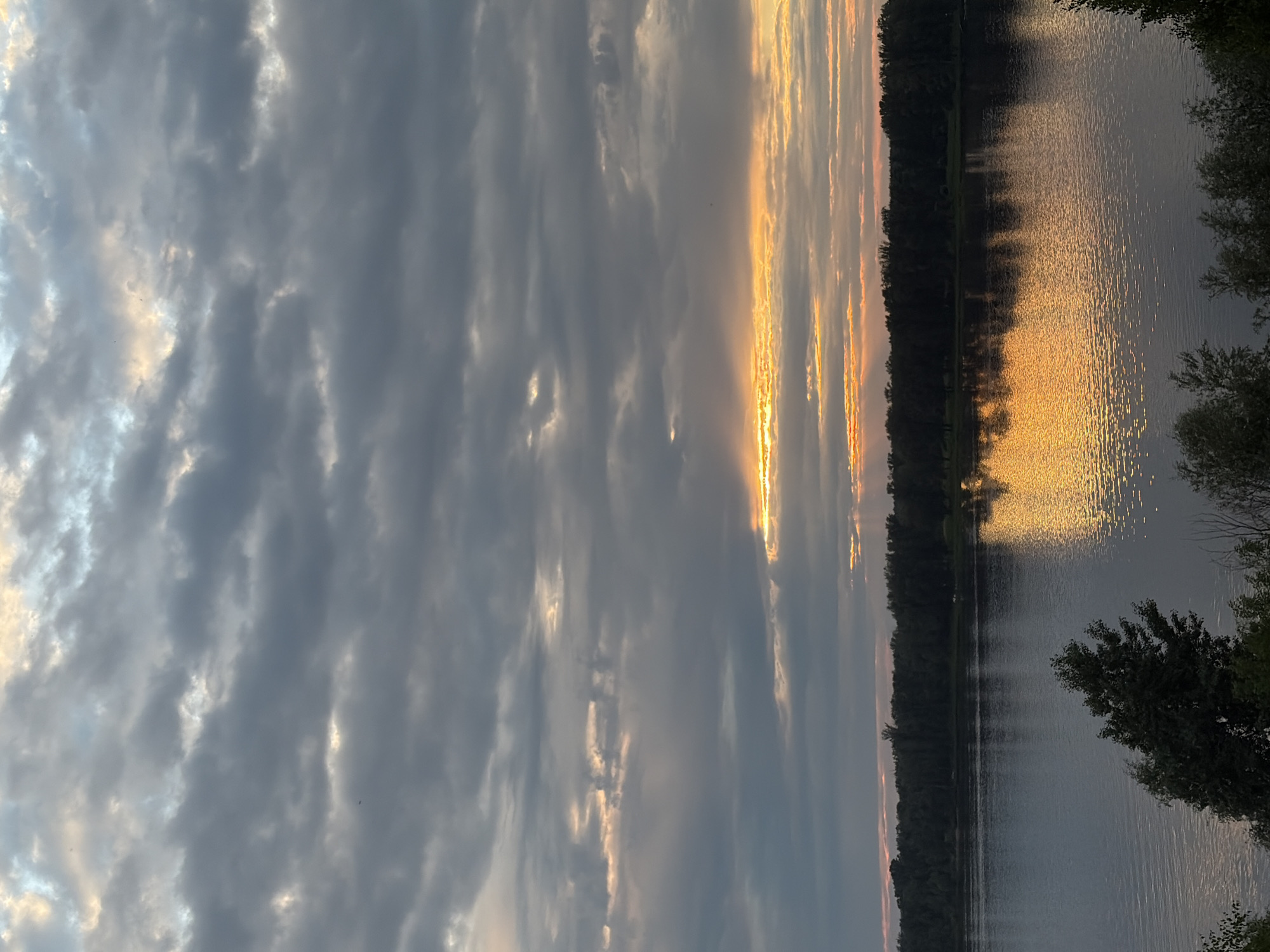 The image depicts a natural landscape at dusk, likely near the village of Olkhovka, Russia, as suggested by the context. The scene is dominated by a wide river that occupies the lower half of the frame. The water is calm, with gentle ripples reflecting the muted colors of the sky. Along the far bank of the river, a dense line of dark trees forms a silhouette against the horizon.
The sky above is a dramatic display of clouds. Heavy, gray clouds cover most of the upper portion of the image, but they are broken by streaks of warm, golden light. These shafts of sunlight suggest that the sun is low on the horizon, possibly just below it, illuminating the undersides of the clouds and casting a warm glow. The light is particularly intense in a band across the middle of the sky, creating a striking contrast with the darker clouds.
In the foreground, the tops of trees and foliage are visible, creating a sense of depth and framing the view of the river and sky. There are no people or animals visible in the scene. The overall atmosphere is serene and contemplative, characteristic of twilight in a natural setting. The specific location within Olkhovka is not identifiable from the image alone, but the presence of a river and dense forest is consistent with the geography of rural areas in Russia.