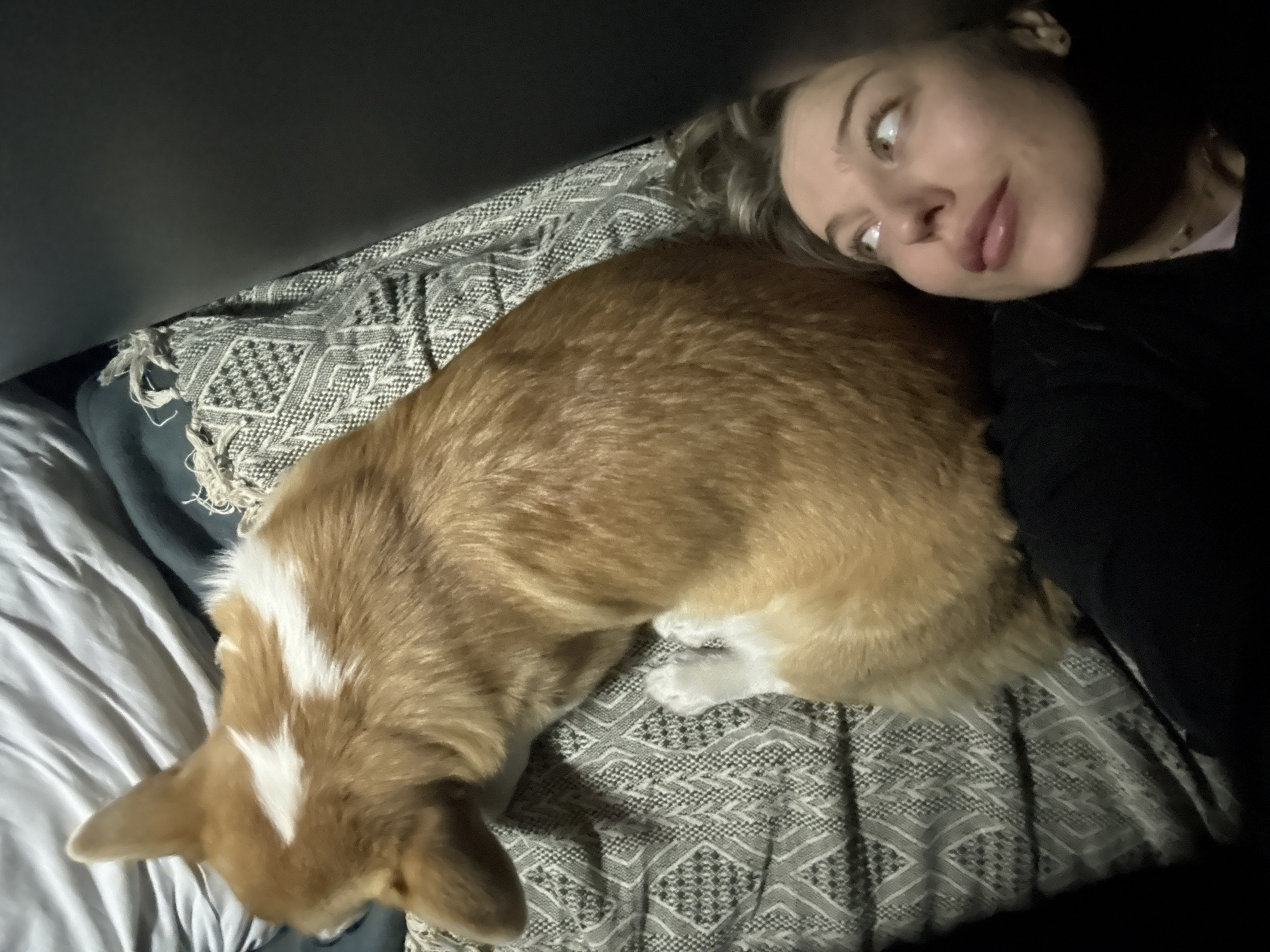 A woman is lying on a bed with a corgi dog. The woman is fair-skinned with blonde hair. She is wearing a black garment. The dog is primarily orange, with white markings on its head and paws. The bed has white sheets and a patterned gray blanket. The image is taken indoors, possibly under a low light condition. There is no location information available in the city of Ага, Russia.