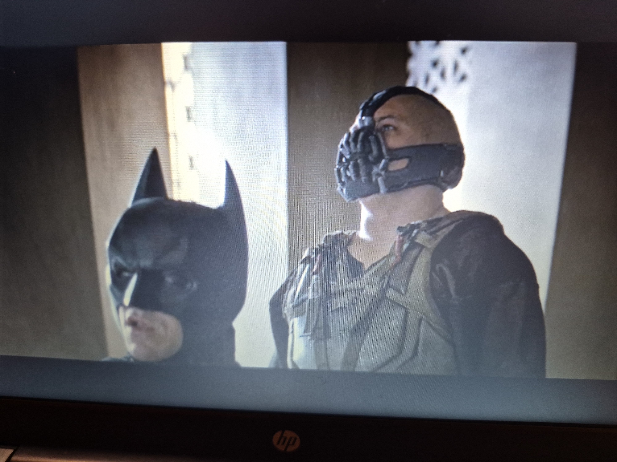 The image shows a screen displaying a scene from a film. On the left is a person wearing a black Batman cowl, with only their eyes and nose visible. To the right of the Batman character is another person wearing a dark, segmented mask that covers their mouth and nose. This individual has short hair and is wearing a tactical vest over dark clothing. They appear to be looking off to the side. The background is dimly lit, with what looks like an indoor setting featuring vertical elements that could be pillars or window frames with intricate patterns. There is no information present to suggest this scene is located in Sandwell, United Kingdom.