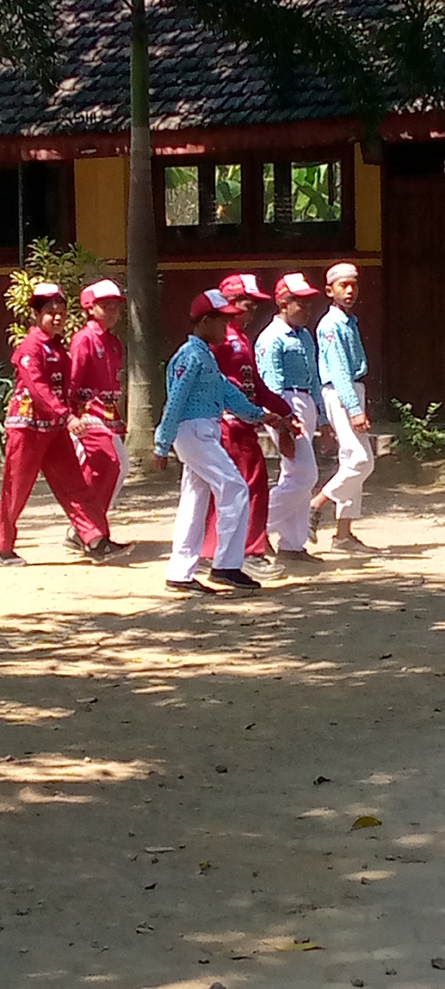 In Pacing, Indonesia, a group of five young boys, likely students, are captured walking outdoors. They are dressed in uniforms, with some wearing red tops and pants, and others in light blue shirts and white pants. All of them are also wearing caps, with red and white variations visible. The boys are walking in a line from left to right across the frame, with some of them interacting with each other, possibly in a parade or procession. The background shows a traditional building with a tiled roof and windows, suggesting an educational institution or cultural site. The ground is covered with dirt and scattered leaves, dappled with sunlight and shadows, indicating an outdoor setting under trees.