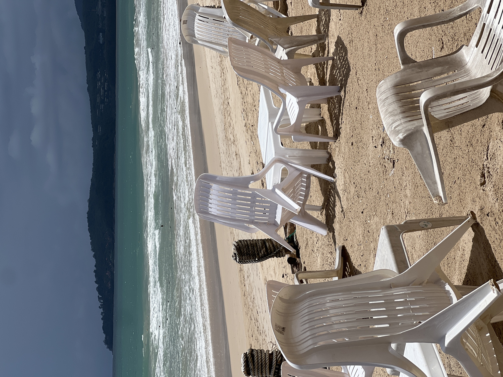 The photograph captures a beach scene in an unknown location. The focus is on a collection of stacked white plastic chairs scattered on the sand. The beach stretches out to the left, where gentle waves roll in towards the shore. In the distance, a dark, tree-covered landmass is visible against a cloudy sky. The overall lighting suggests a sunny day, casting shadows around the chairs.