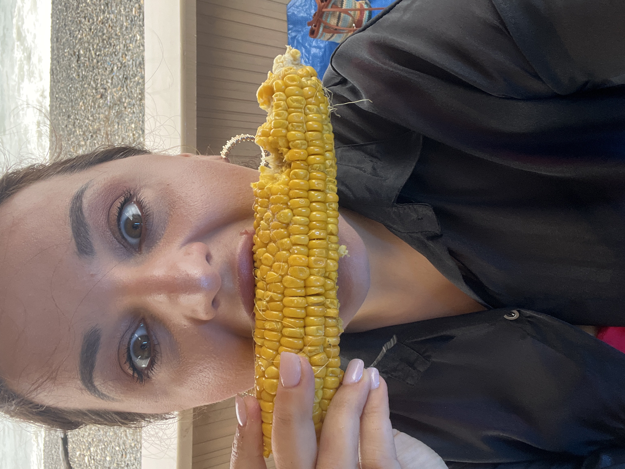 In this close-up, waist-up shot, a woman with dark hair and makeup is holding a partially eaten corn on the cob to her mouth. She is wearing a black button-up shirt, and large hoop earrings. Her nails are painted a pale pink. The background appears to be a beach or waterfront scene, with a rocky shore and water visible. The lighting suggests it is daytime. The woman's expression is one of surprise or excitement.