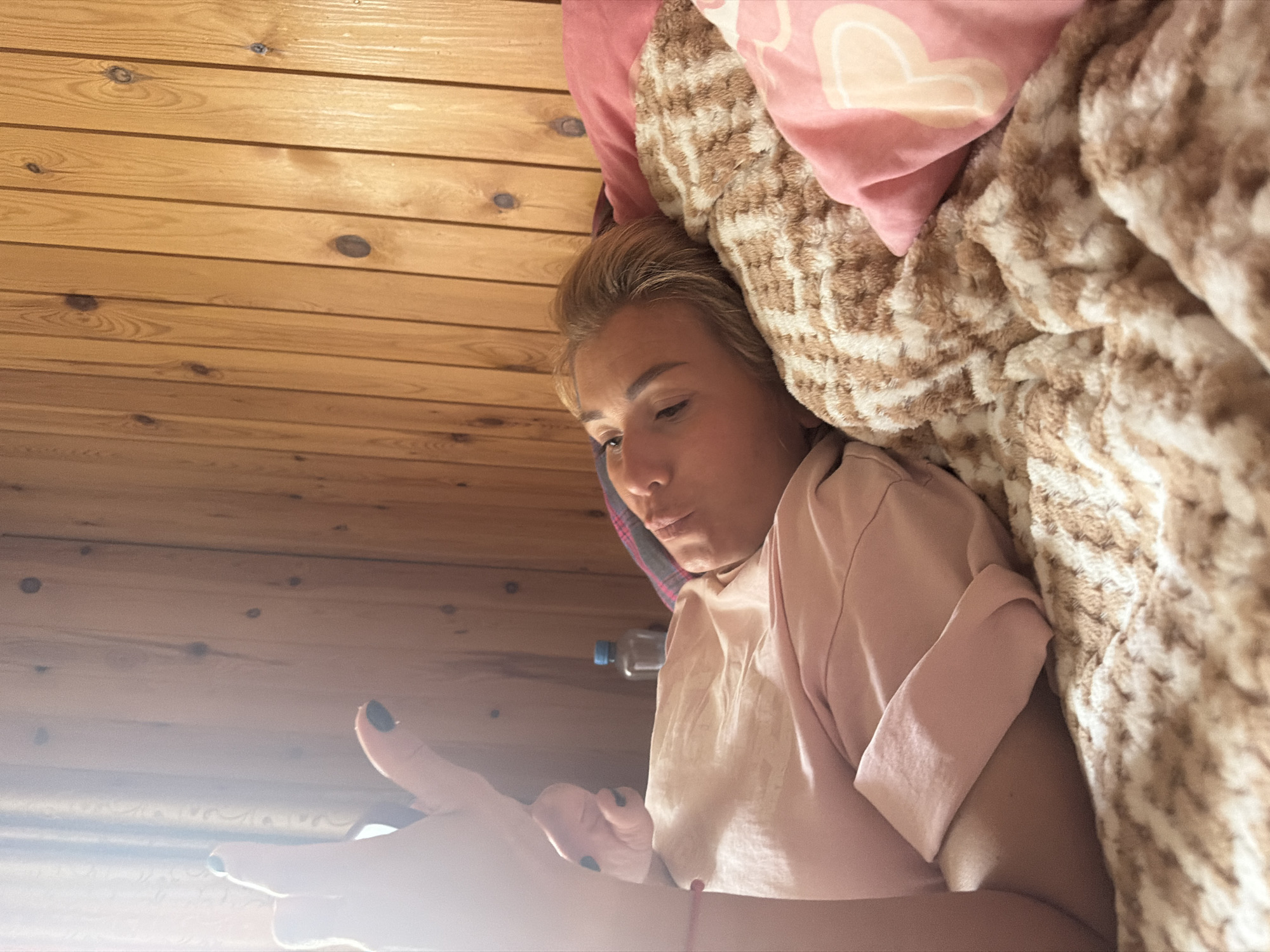 An overhead medium shot shows a young adult with blonde hair lying on their side in bed, facing away from the camera and looking to the left. The subject is wearing a light pink t-shirt and is lying on a bed with a brown and white patterned comforter. To the right of the subject is a pink pillow with a light pink "P" logo. The background consists of a wooden paneled wall. To the left of the frame, the subject's left arm is raised, with their hand in focus, displaying black nail polish. A faint pink bracelet is visible on the wrist. A bottle of water is present on the bedside table behind the subject. The scene suggests a relaxed, indoor setting within a room that has a rustic aesthetic, likely a bedroom or guest room. The lighting is soft and diffused, possibly indicating natural light entering from a window, which casts a gentle glow on the subject and the immediate surroundings. The overall impression is one of casual comfort.