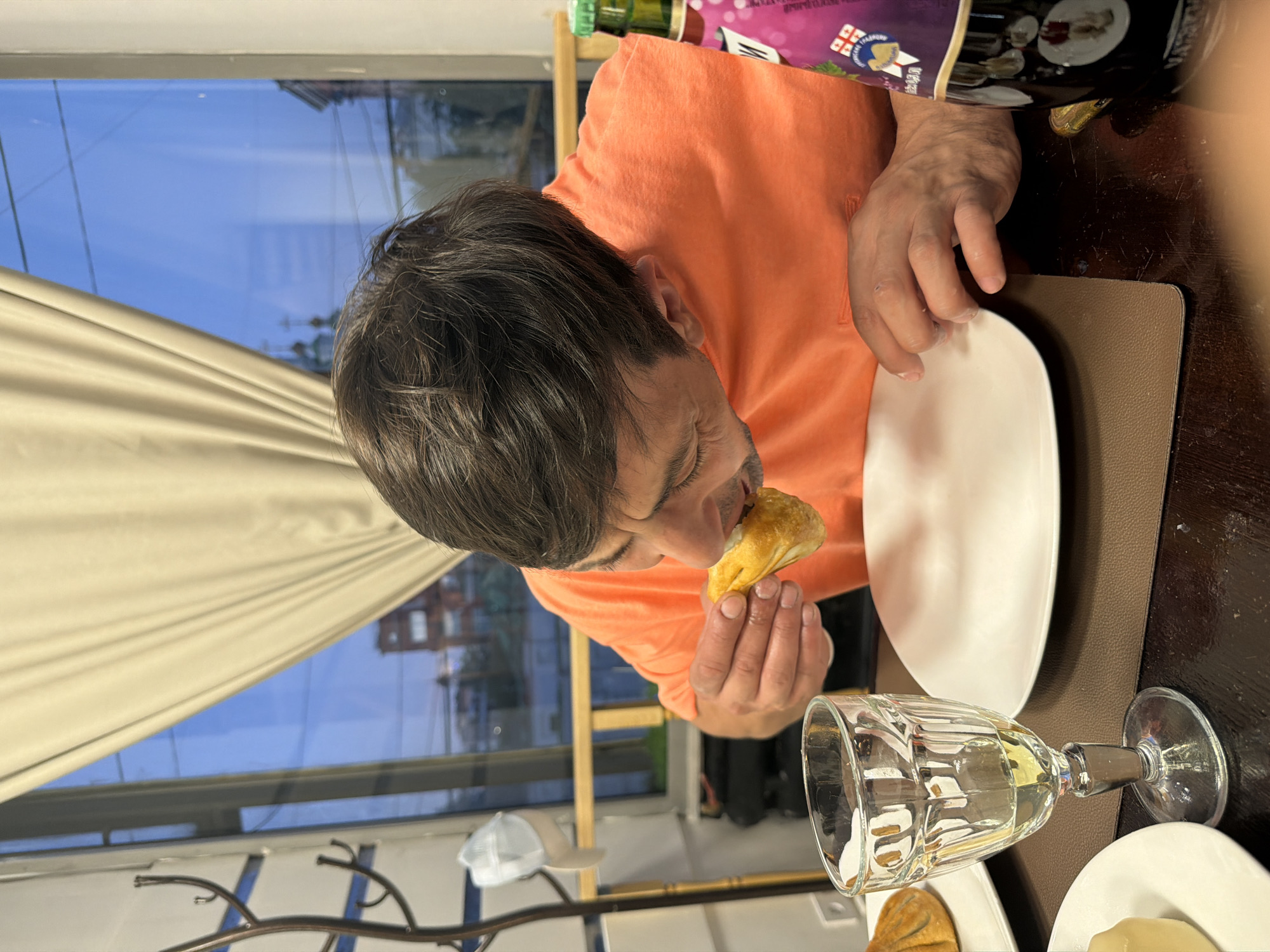 An adult male is captured mid-meal in an indoor setting, seated at a dark-colored table. He has dark hair and is wearing a bright orange short-sleeved shirt. He is shown holding a golden-brown, pastry-like food item, possibly a dumpling or a baked good, with both hands and bringing it to his mouth to eat. His expression suggests focus on the food.
On the table in front of him, there are white rectangular plates, one of which holds another similar food item. A clear glass with a stem, containing a light-colored liquid (possibly white wine or water), is also visible on the table. A rectangular light brown placemat is beneath some of the dishes.
In the upper background, a large window is visible, through which a bright blue sky can be seen, indicating it is daytime. Parts of light beige curtains are drawn back, revealing reflections of buildings or structures outside the window. To the right, a partially visible bottle or packaging with a purple label and a green cap is present. Text on the label includes "GEORGIAN NATU..." and a design resembling the Georgian national flag. Some other white text is also visible but illegible. The overall scene depicts a casual meal being enjoyed indoors.
