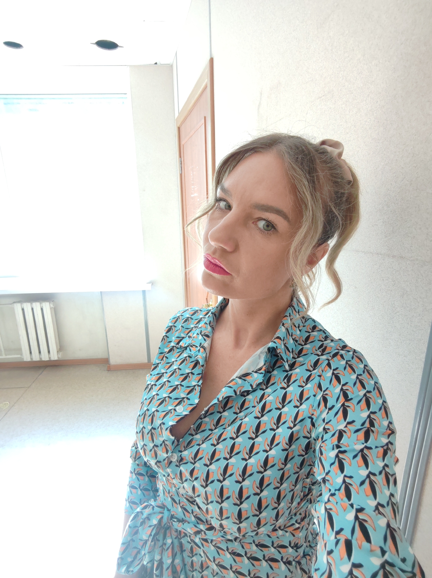 A woman with blonde hair pulled back into a messy bun, wearing a blue blouse with a leaf pattern, is posing for a selfie in what appears to be an indoor hallway. The walls are a light beige or off-white color. There is a radiator visible against a white wall, and a doorframe is also partially visible. The location is in the city of Unknown, Unknown.