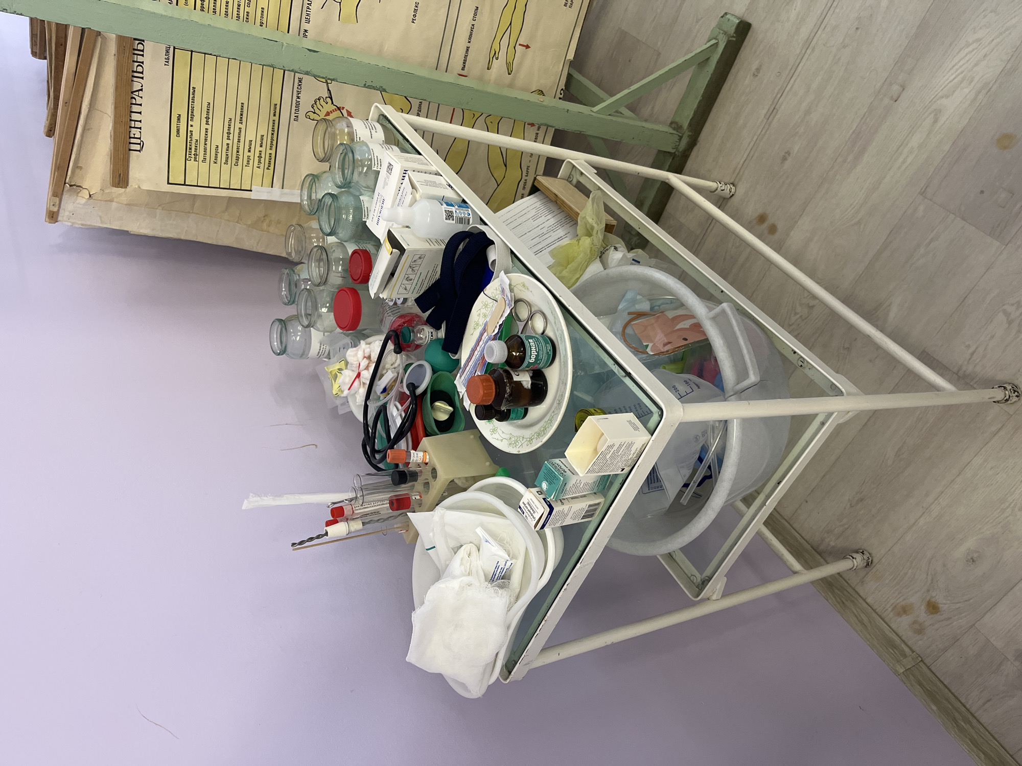 This image depicts an indoor medical setting, likely a clinic or educational facility, located in Chaikovsky, Russia. A white, two-tiered metal medical trolley with glass shelves stands in the foreground, laden with various medical supplies.
The upper shelf of the trolley holds numerous glass jars, some with red lids, alongside bottles of liquids, including one prominently labeled "борная" (boric acid). Also on this shelf are blue medical gloves, a stethoscope, several small boxes of unknown contents, and a white plate containing medical instruments like forceps and scissors, surrounded by smaller bottles. The lower shelf contains a white plastic basin, a container with gauze and cotton swabs, more small boxes of supplies, and test tubes with syringes in a rack. The arrangement suggests an active medical or training environment.
Behind the trolley, a large, yellowed medical chart or poster in Cyrillic (Russian) text is displayed on a green metal stand. Visible headings on the chart include "ЦЕНТРАЛЬНЫ" (CENTRAL), "ТАБЛИЦ" (TABLE), "СИМПТОМЫ" (SYMPTOMS), "Сухожильные и периостальные рефлексы" (Tendon and periosteal reflexes), "Патологические рефлексы" (Pathological reflexes), "Клонусы" (Clonus), and "ВЫЯВЛЕНИЕ КЛОНУСА СТОПЫ" (DETECTION OF FOOT CLONUS). It details various neurological signs and reflexes.
The room features light purple walls and a light-colored wooden floor, with some visible stains. The overall appearance, including the style of equipment and chart, suggests a functional and possibly older facility. No people are visible, indicating the scene is captured either before, between, or after medical activities such as examinations, procedures, or teaching sessions.