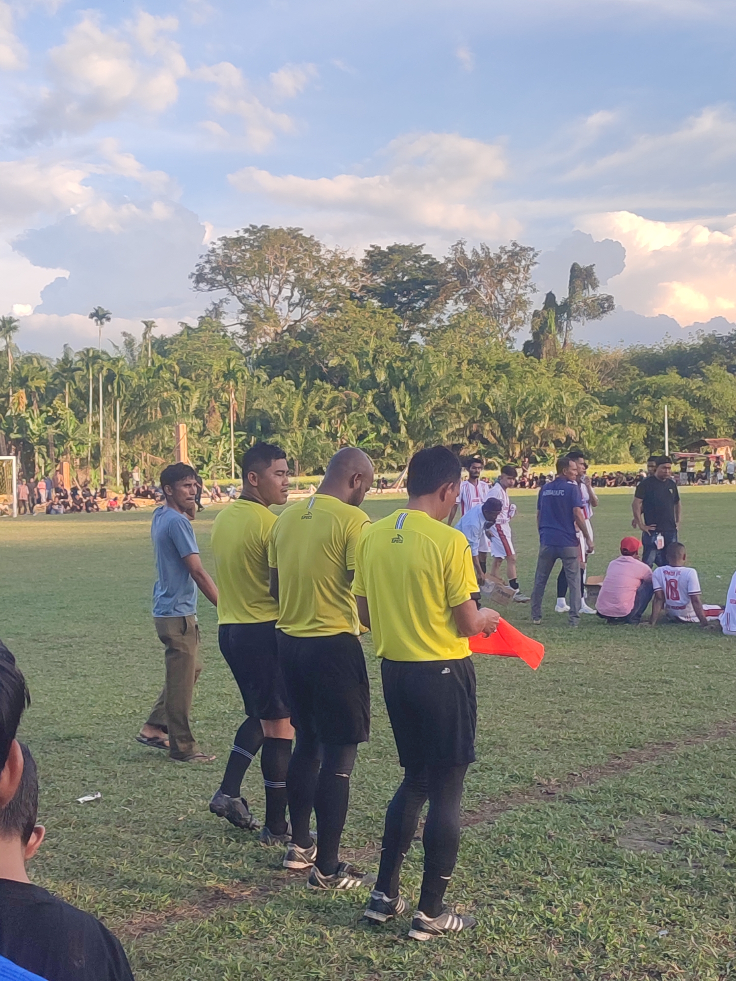 A daytime outdoor scene on a grassy field in Babahlueng, Indonesia, features a group of people, presumably involved in a soccer match. In the foreground, four individuals, wearing yellow soccer jerseys and black shorts with black socks, are seen from the back. The person farthest to the right is holding an orange flag. Another man to the left of the referees is looking into the distance. A crowd of people is visible in the background, watching the game. Trees line the perimeter of the field. The sky above is partly cloudy.