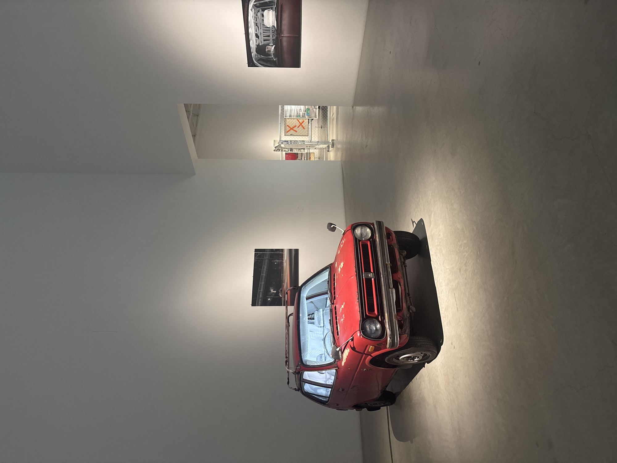 This image captures an art installation within a modern, minimalist gallery space, likely in Kanazawa, Japan. The central subject is a vintage, red two-door car, possibly a Honda Life or similar model, positioned on a light concrete floor. The car is heavily worn, with visible rust and chipped paint, contributing to its artistic aesthetic. Its interior, dimly lit with a cool blue light, adds an ethereal quality.
On the white wall to the left of the car, two framed photographs are displayed. The upper photograph shows a close-up detail of a car's front grille and headlight, rendered in black and white, possibly hinting at the car's origins or a broader automotive theme. The lower photograph is darker, depicting what appears to be a blurred nocturnal scene, perhaps a city street or highway view from a moving vehicle.
In the background, through a rectangular opening in the wall, another area of the gallery is visible. This section features industrial-style metal barriers or scaffolding. On a white surface or sign behind these barriers, two large red 'X's are prominently displayed. The overall lighting is artificial and subtle, casting soft shadows that highlight the car's form and the textures of the space. The environment is clean and stark, emphasizing the artworks as focal points.