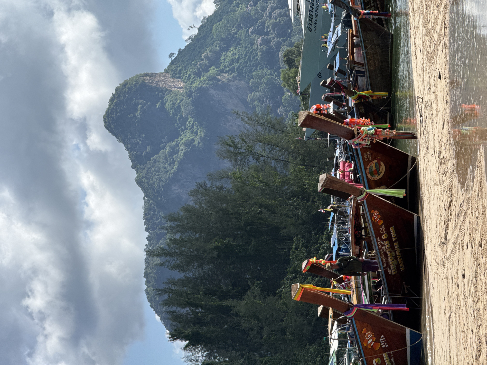 The image shows a beach in Ban Klong Haeng, Thailand, with several longtail boats docked. The boats are wooden and have colorful fabric decorations. In the background, there is a large mountain covered in lush green trees. The sky is cloudy with patches of blue. A person in a green shirt and dark pants is standing on one of the boats. A larger boat, possibly a ferry, is visible behind the longtail boats, with text that reads "AO NANG PRINCE". The beach is sandy and there are tire tracks visible on it. The water is calm and reflects the colors of the boats and the sky.