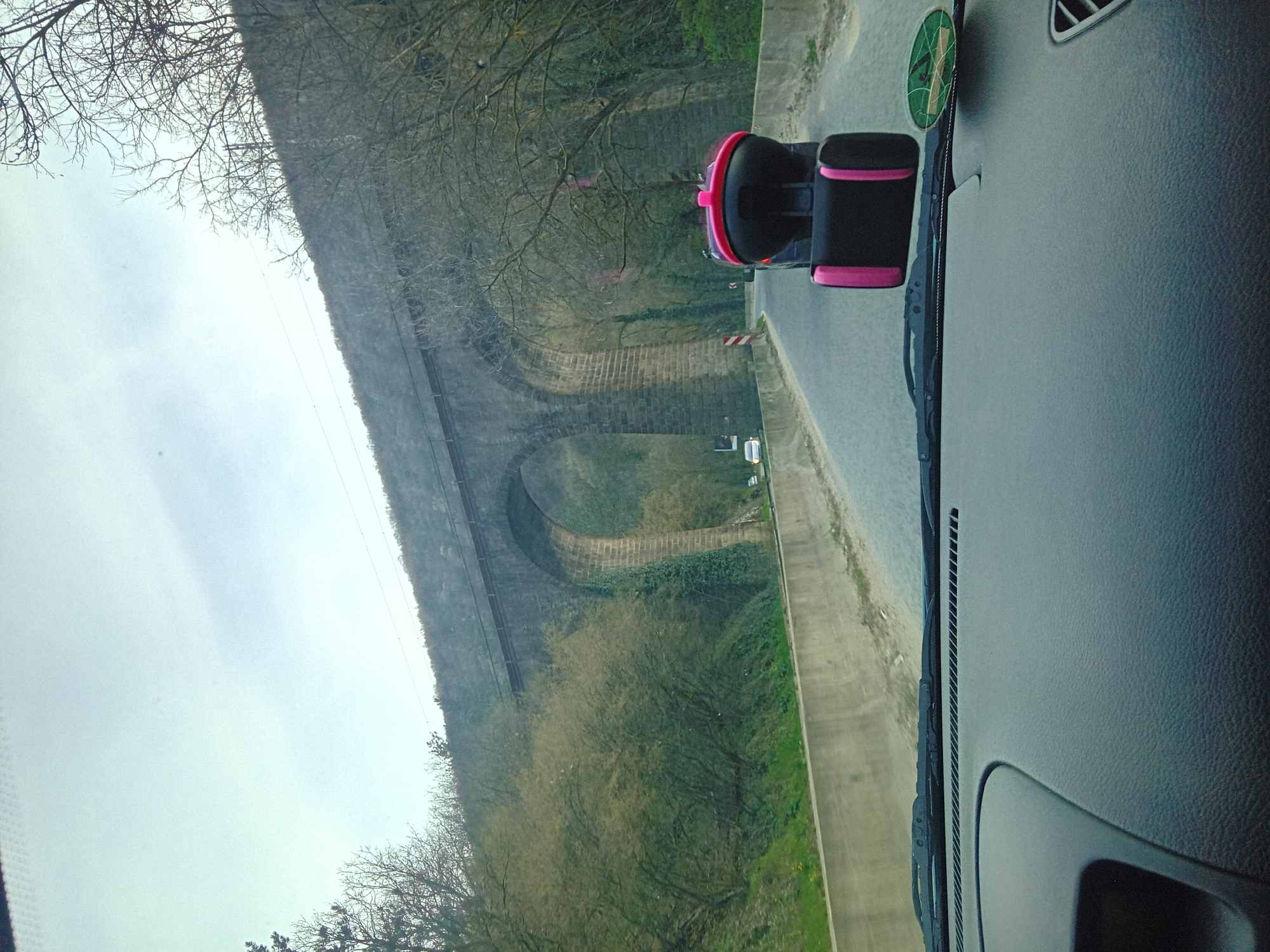 The image, taken from inside a vehicle, shows a viaduct running parallel to a road. The viaduct appears to be made of stone or brick and has a series of arches. The vehicle's dashboard and a phone holder are visible in the foreground. Beyond the road, there are trees and vegetation along the viaduct's base. The sky is overcast. The location is unknown, as is the city.