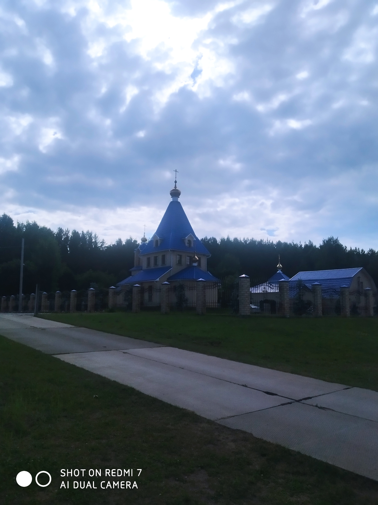 An eye-level outdoor shot depicts an Eastern Orthodox church complex located in Chaikovsky, Russia, under an overcast sky. In the foreground, a paved pathway with visible seams and cracks leads into the midground, bordered by green grass.
The central subject is a light-colored church building featuring prominent blue, multi-tiered domed roofs, capped with golden onion domes and crosses. To the right of the main structure, a smaller building with a similar blue roof and a modest golden dome is visible. Both buildings are enclosed by a fence composed of sturdy brick pillars and dark metal railings.
A dense, dark green forest forms the backdrop behind the church complex, suggesting a secluded or suburban setting. The sky is dominated by thick, light gray clouds, with brighter patches indicating diffused daylight. The overall atmosphere is serene and quiet, with no people or overt activities visible. A digital watermark in the lower-left corner reads "SHOT ON REDMI 7 AI DUAL CAMERA".